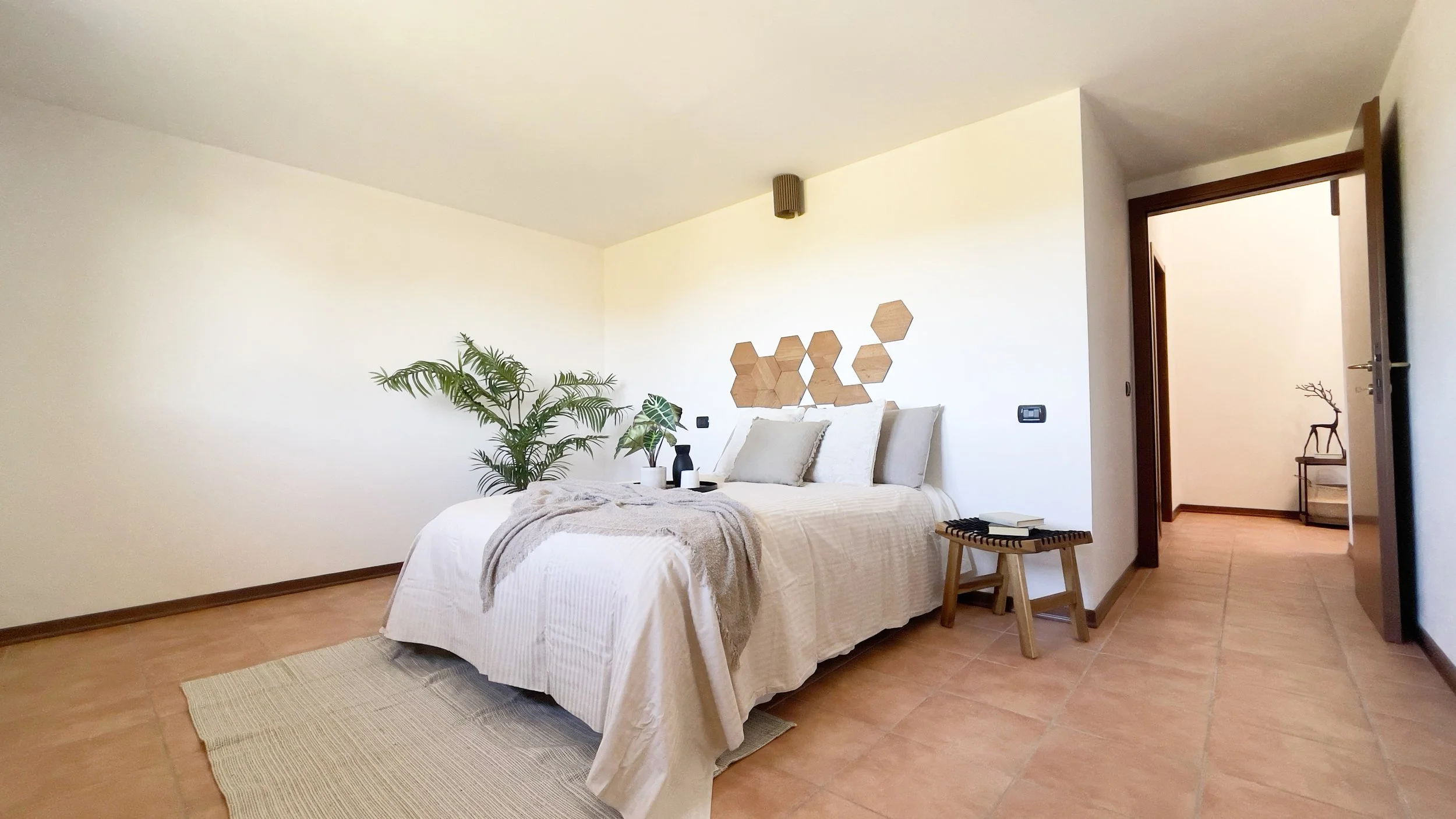 Camera da letto moderna con letto matrimoniale e decorazioni sulle pareti, pavimento in piastrelle, piante verdi e uno sgabello con libri accanto al letto.