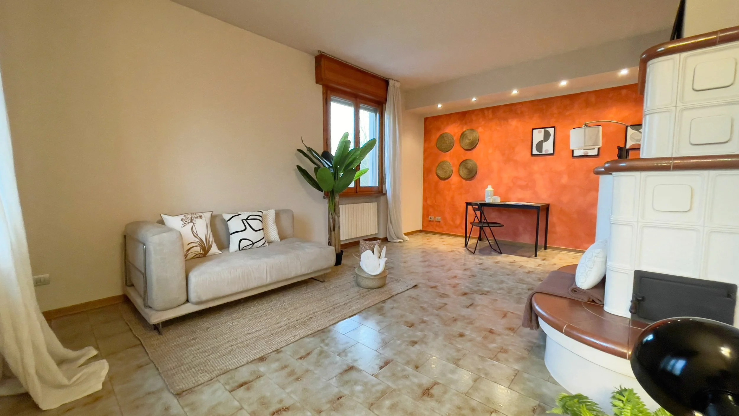 Soggiorno luminoso con divano beige, piante verdi, parete arancione con decorazioni, tavolo con sedia, tappeto e stufa bianca.
