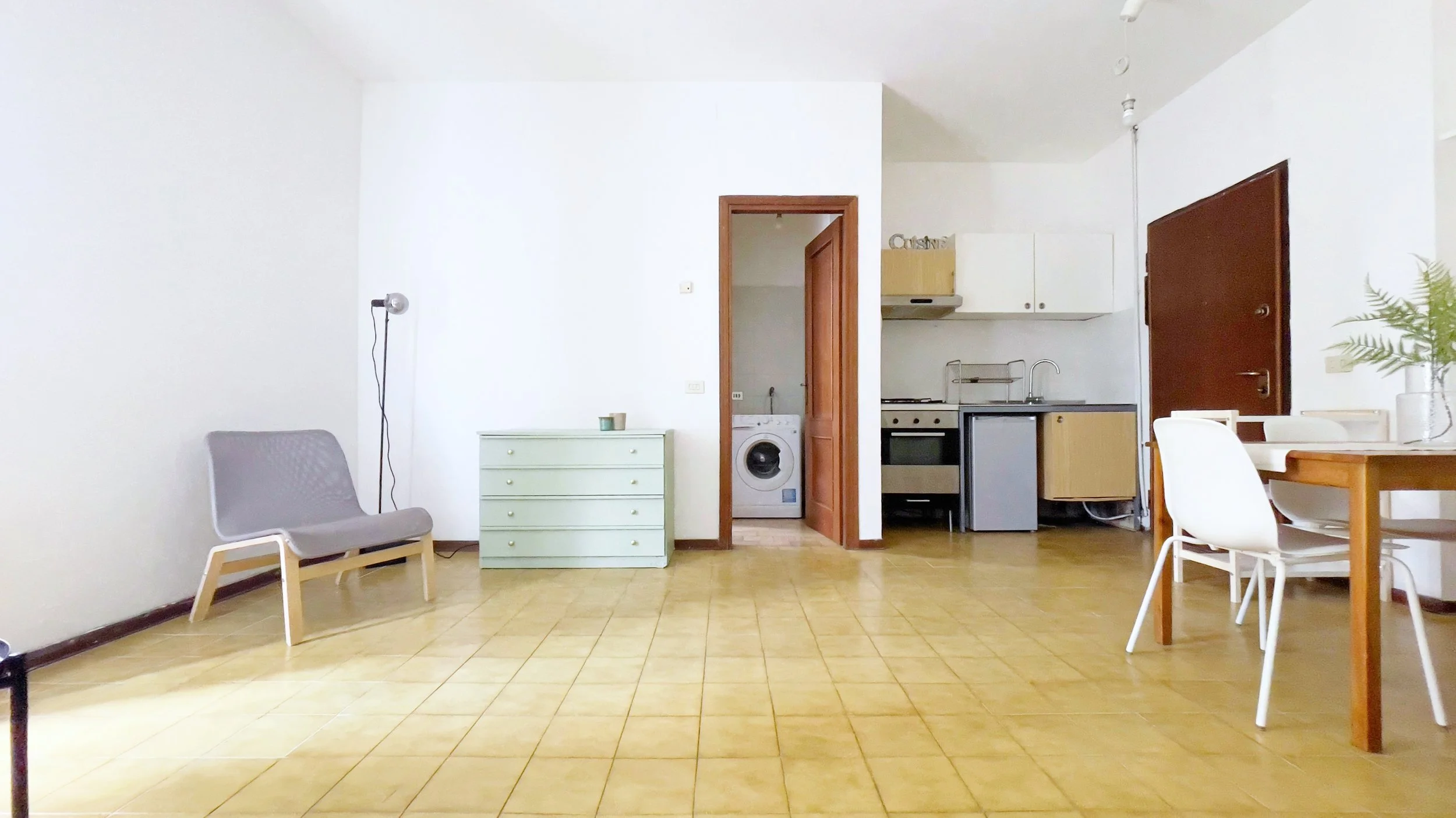 Interno di un appartamento con pavimento in piastrelle gialle, una sedia grigia, una cassettiera verde, un tavolo con sedie bianche, una cucina con forno e lavandino, e una lavatrice visibile attraverso una porta aperta.