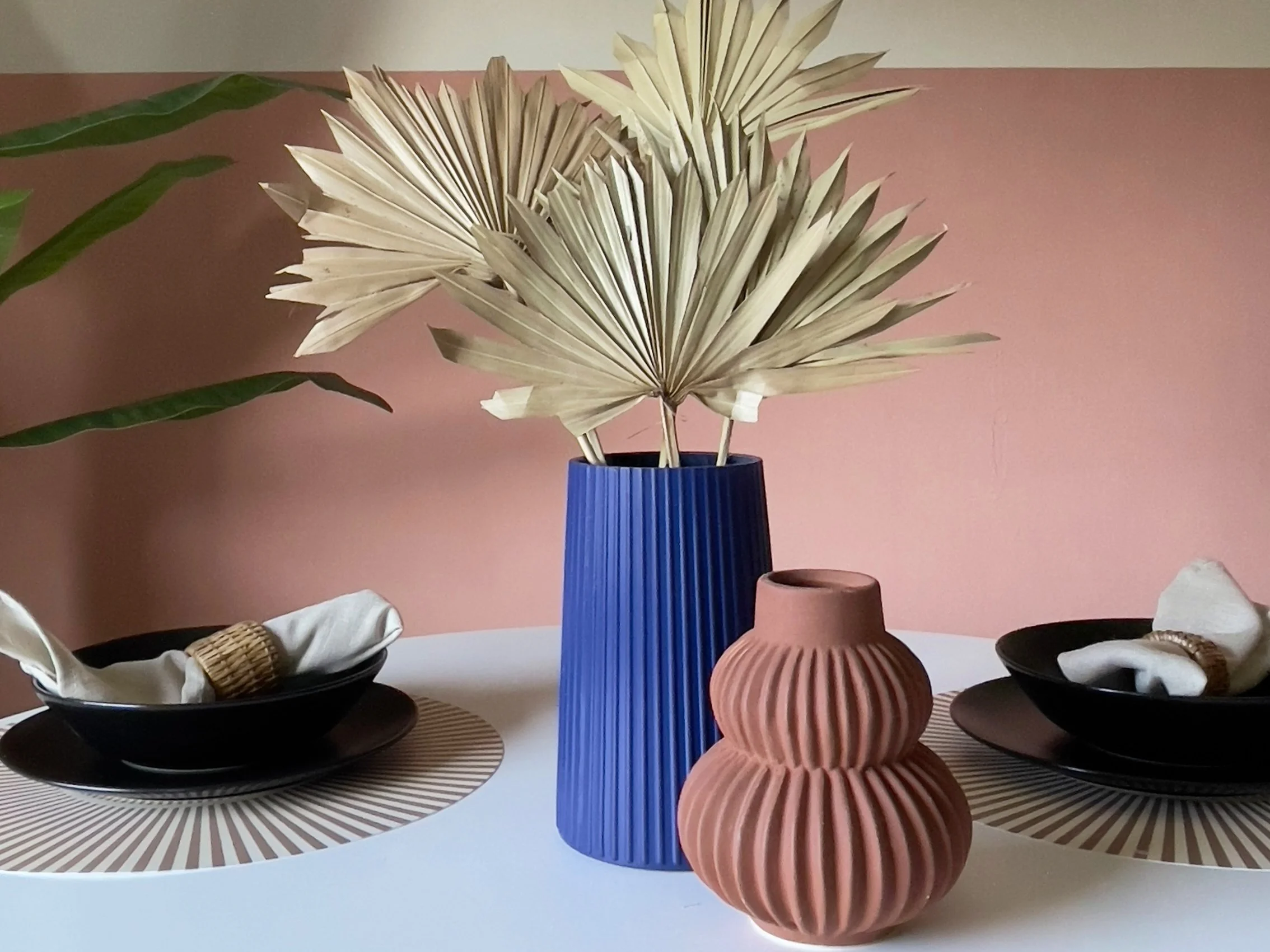 Tavolo apparecchiato con piatti neri, tovaglioli chiari arrotolati in anelli di vimini, vaso blu con foglie essiccate di palma, piccolo vaso decorativo marrone, sfondo rosa e pianta verde a sinistra.