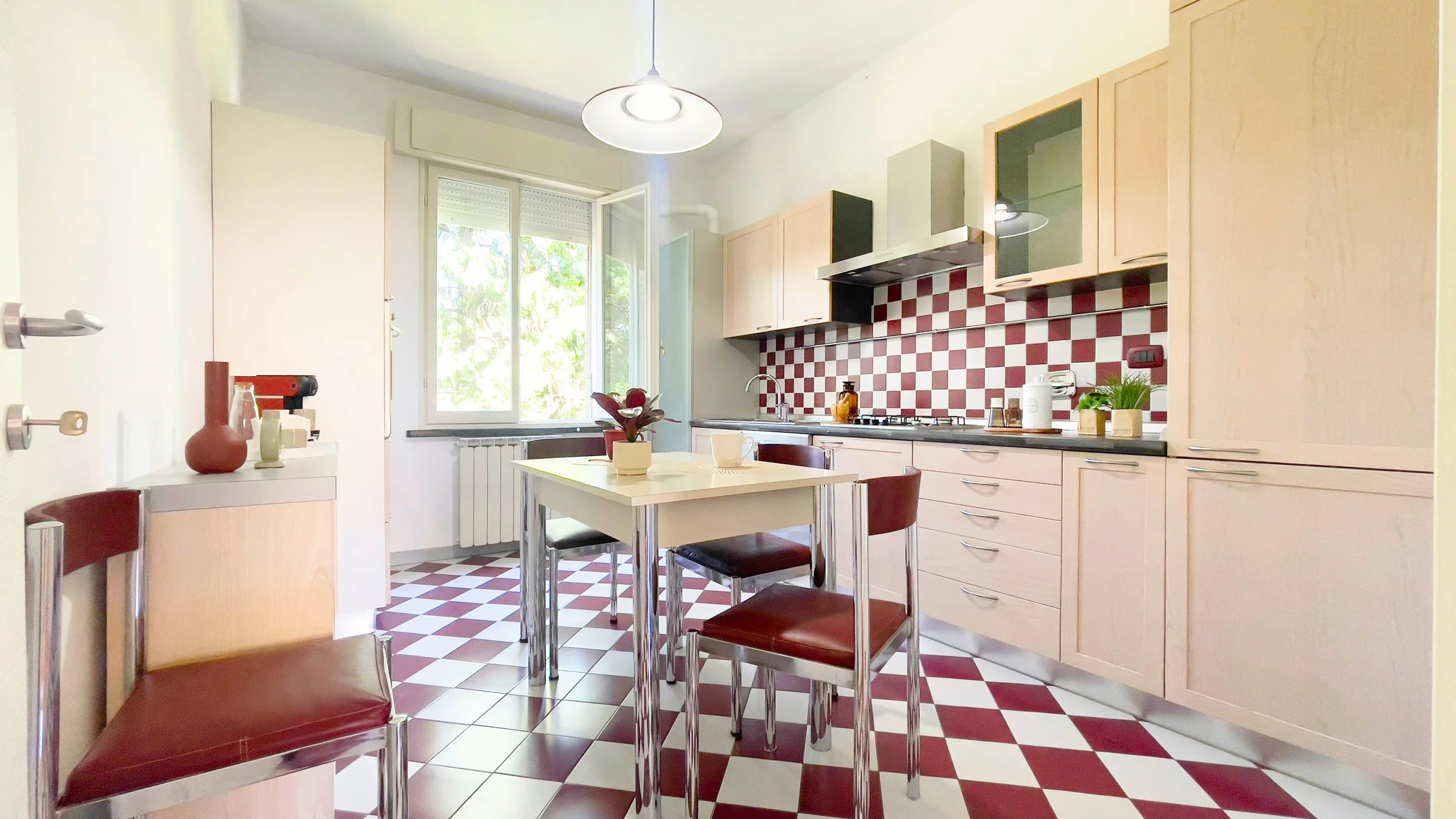 Cucina moderna con mobili in legno chiaro, pavimento a scacchi rossi e bianchi, tavolo con sedie, pianta decorativa, finestra con vista su alberi.
