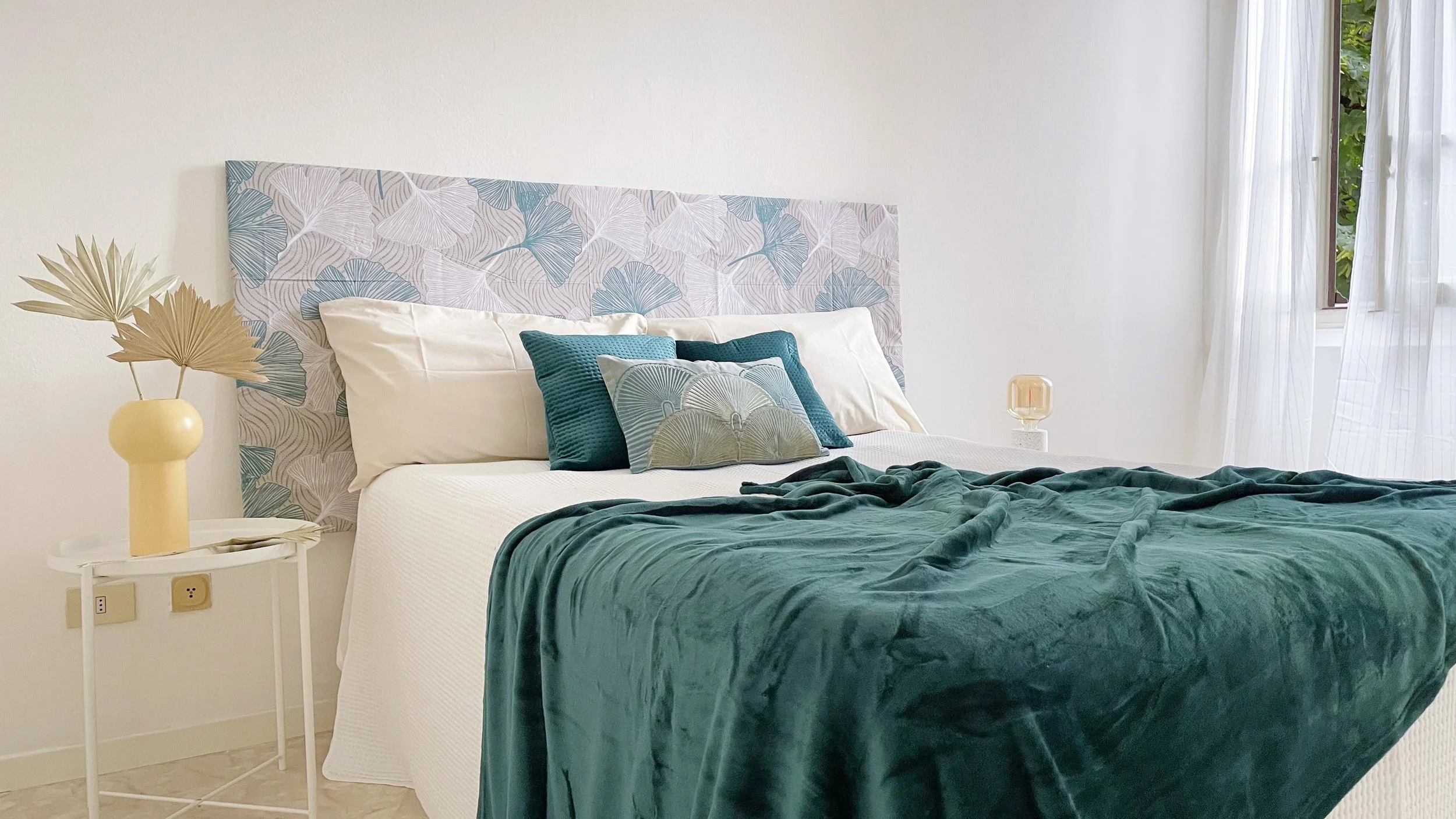 Camera da letto moderna con letto matrimoniale, cuscini decorativi e copriletto verde scuro; testiera con motivo a foglie. Tavolino laterale con vaso e foglie decorative. Finestra con tende bianche.