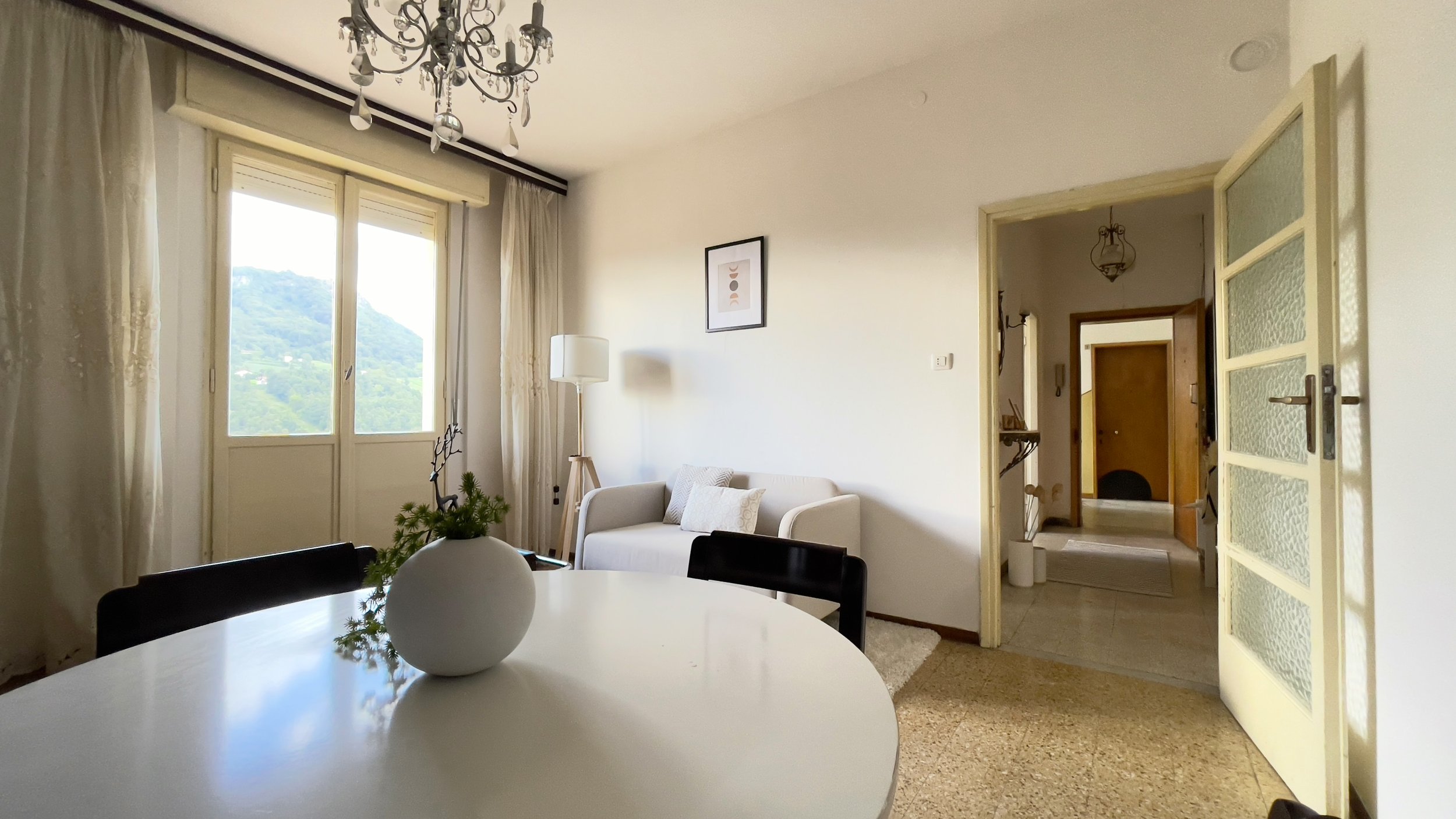 Interno di un soggiorno luminoso con tavolo bianco, sedia nera, vaso decorativo, divano beige, e lampada da terra. Vista di una montagna dalla finestra con tende bianche.