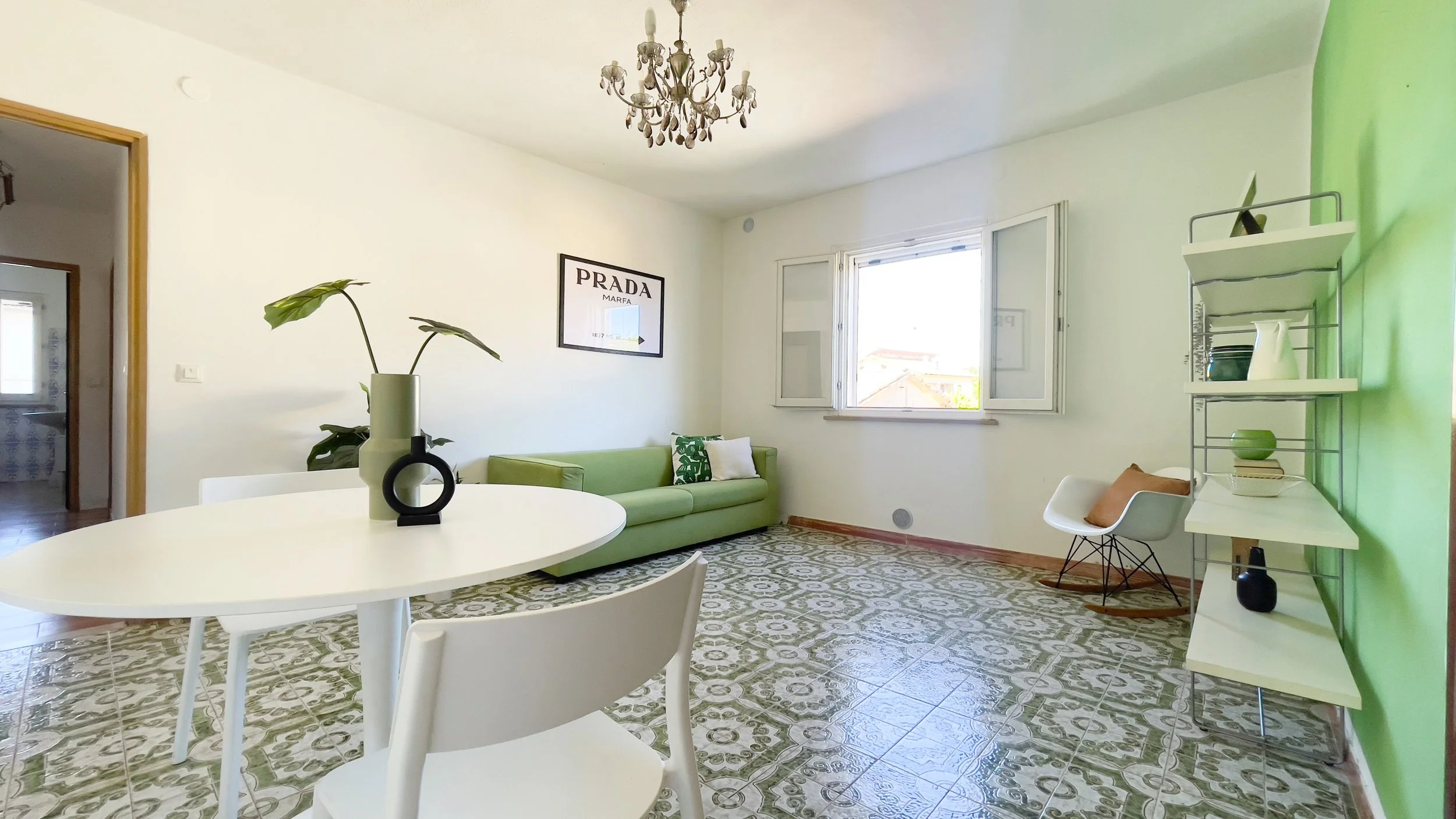 Soggiorno con pareti bianche e verdi, pavimento decorato, tavolo bianco con vaso e sedie abbinate, divano verde, scaffale con oggetti decorativi, e finestra aperta che illumina la stanza.