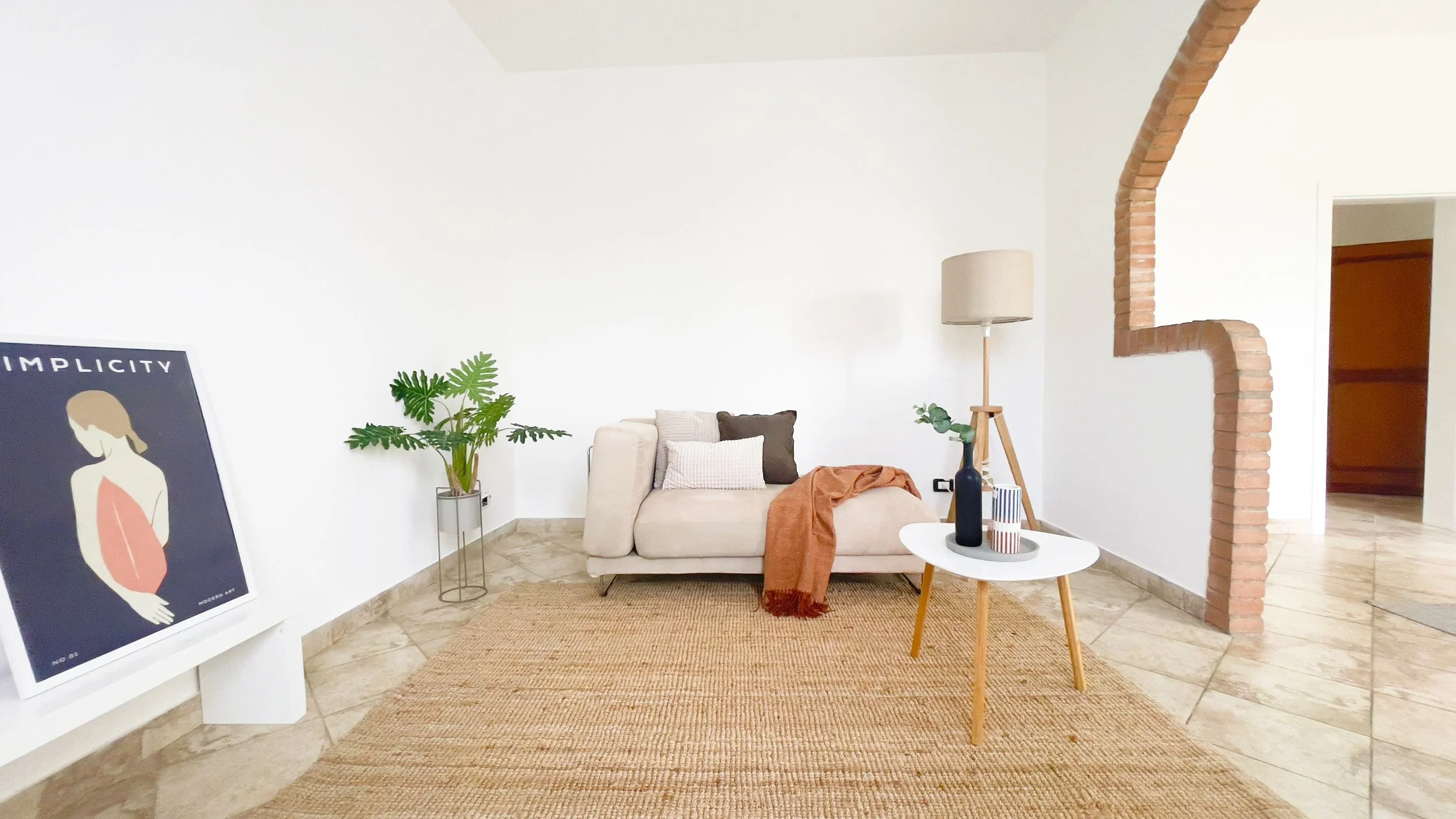 Interno di un salotto moderno con divano beige, cuscini e coperta arancione. Tavolino bianco con decorazioni, lampada a stelo, pianta in vaso e quadro "Simplicity". Parete bianca con dettaglio di arco in mattoni.