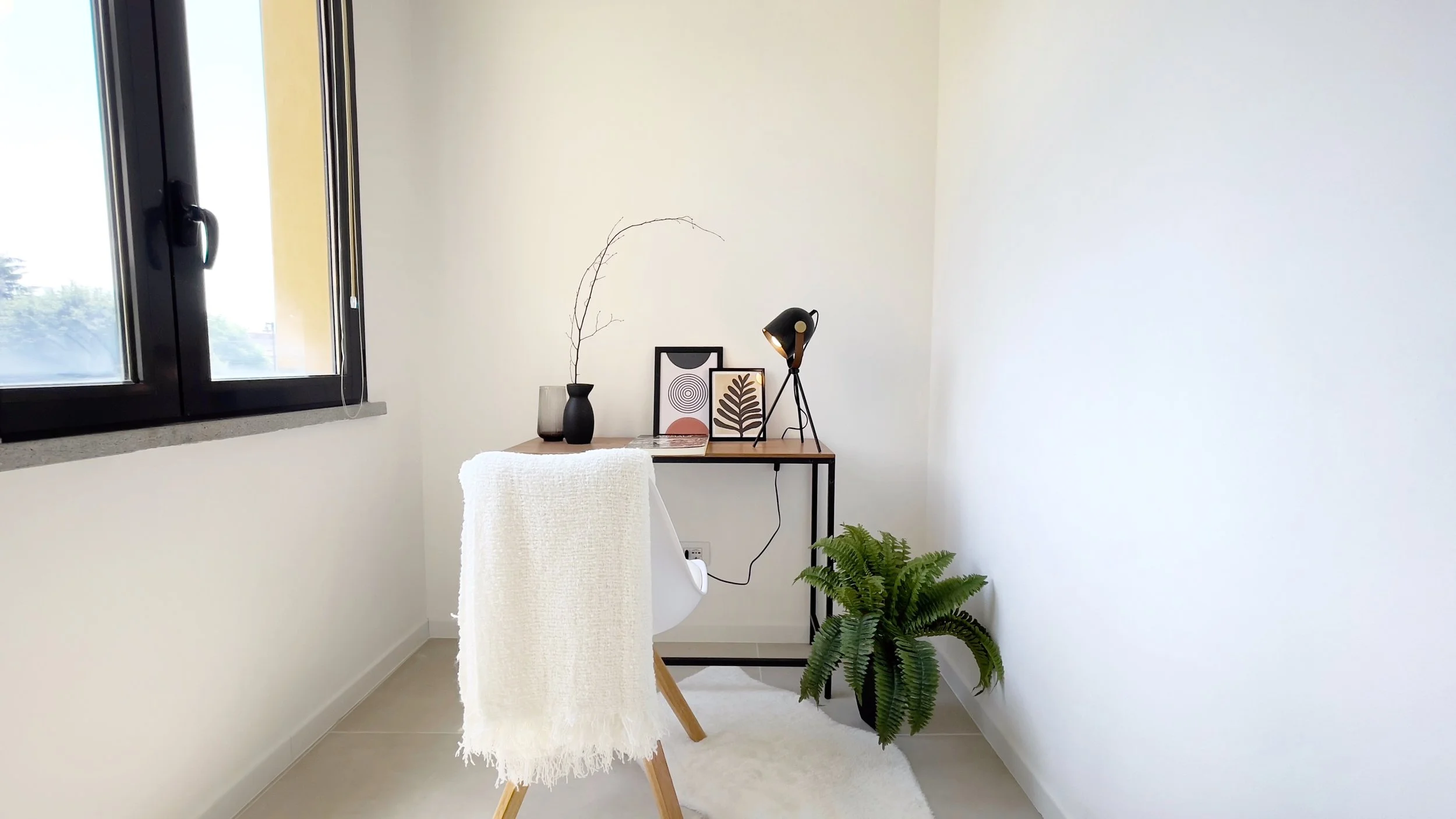 Ufficio minimalista con una scrivania in legno, una sedia bianca con coperta bianca, lampada da tavolo nera, decorazioni artistiche, pianta verde e una finestra aperta sulla sinistra.