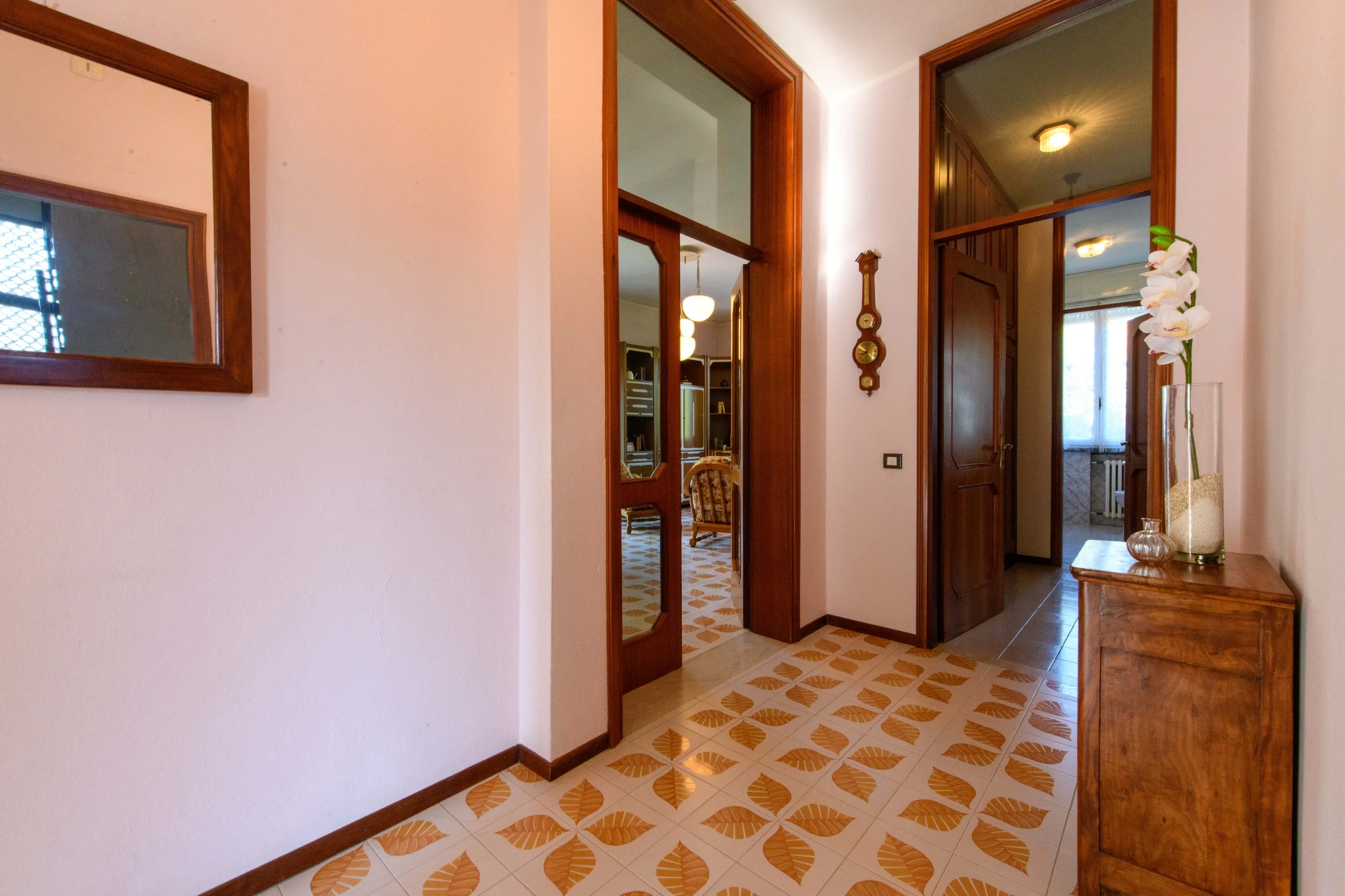 Ingresso di una casa con pavimento a piastrelle con motivi a foglia, specchio con cornice in legno, mobile in legno con vaso di fiori, orologio da parete e porta in legno aperta verso un'altra stanza.