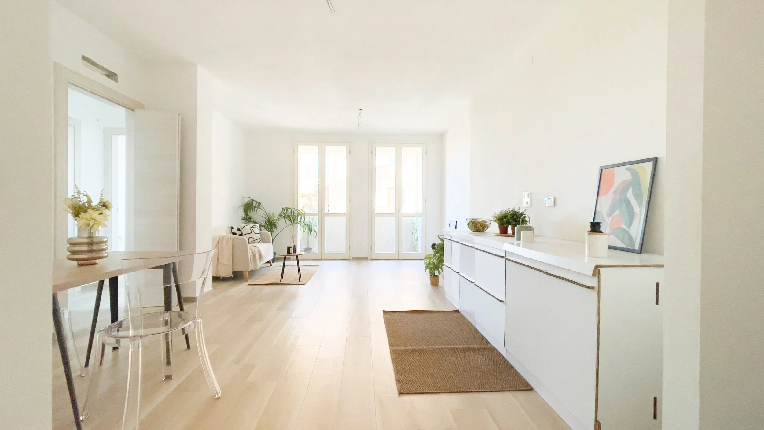 Interno di un salotto moderno con mobili bianchi, tavolo in legno con sedia trasparente, divano, piante e ampie finestre luminose.