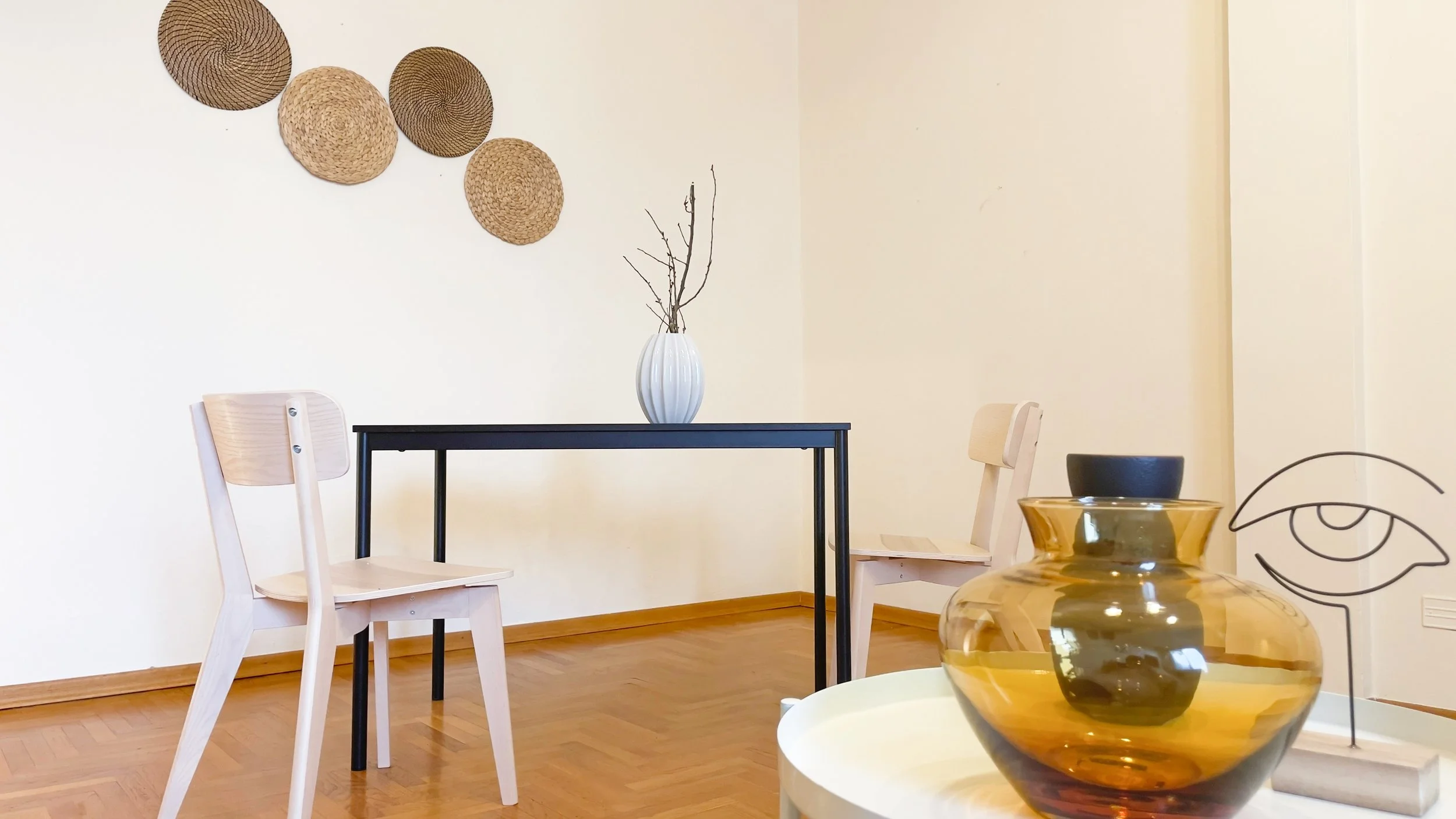 Interno minimalista con tavolo nero, due sedie in legno chiaro, vasi decorativi e oggetti d'arte su un tavolino, parete decorata con piatti in vimini.