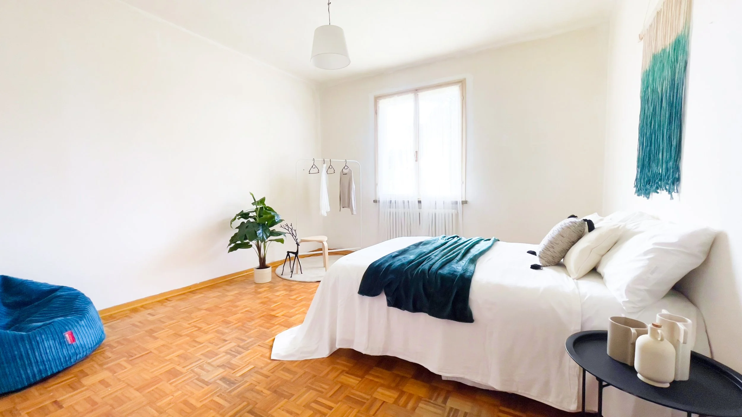 Camera da letto minimalista con letto bianco, tappeto in legno, pianta decorativa, sacco a pelo blu, appendiabiti con abbigliamento e finestra luminosa.