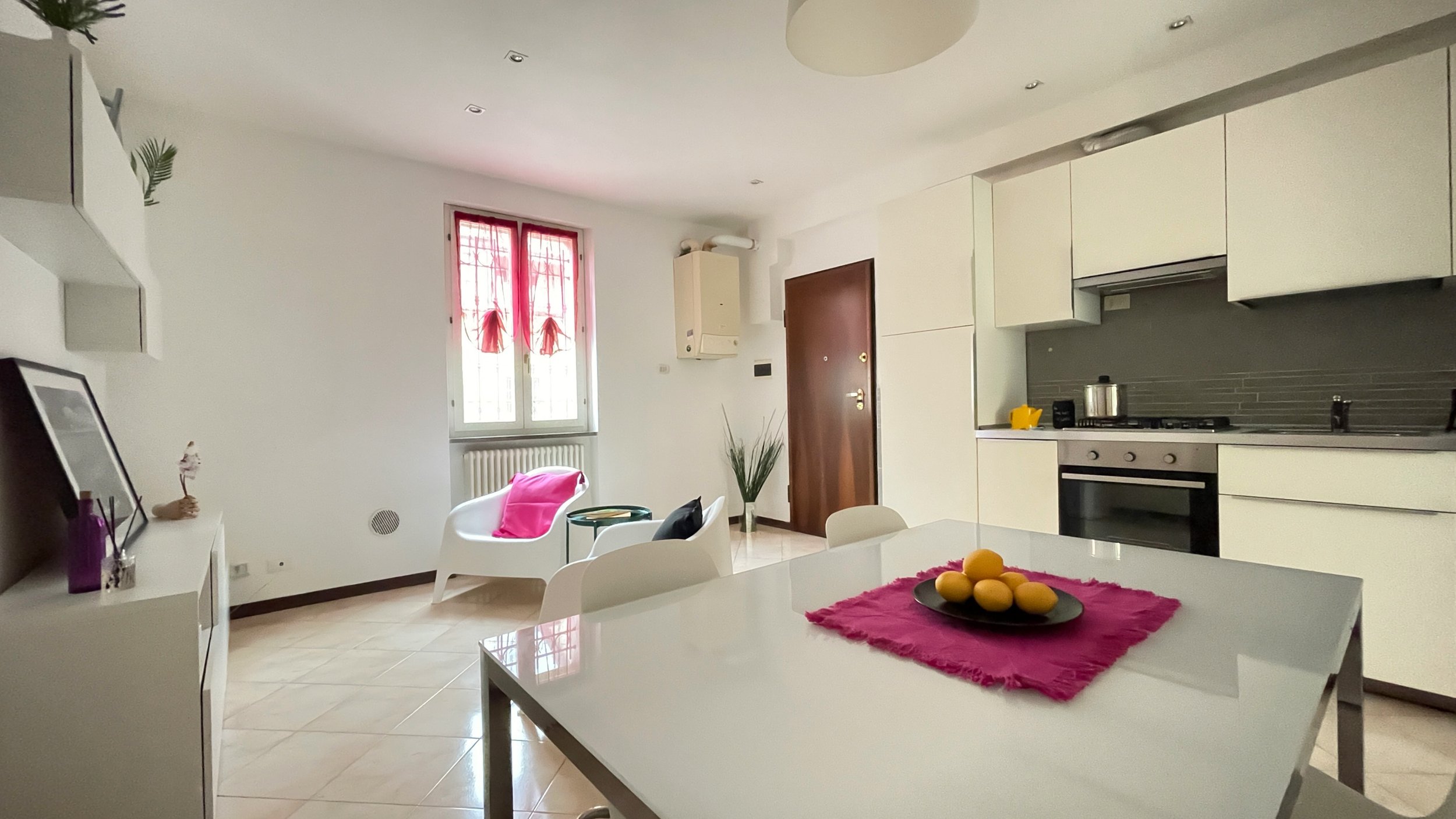 Soggiorno moderno con cucina a vista, tavolo bianco con frutta, sedia e poltrona con cuscino rosa, finestra con tende rosse, pareti bianche, e decorazioni minimaliste.