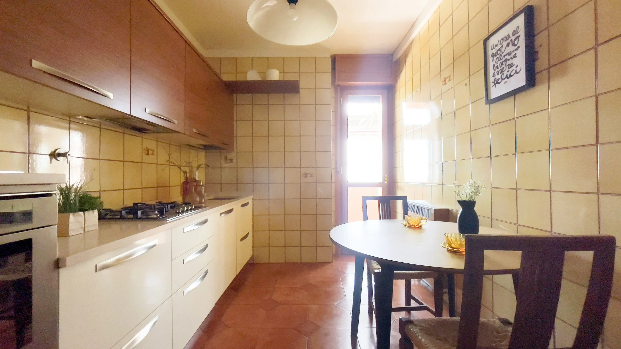 Cucina moderna con mobili chiari e luci soffuse, tavolo rotondo con sedie e vaso decorativo, piastrelle color crema sulle pareti.