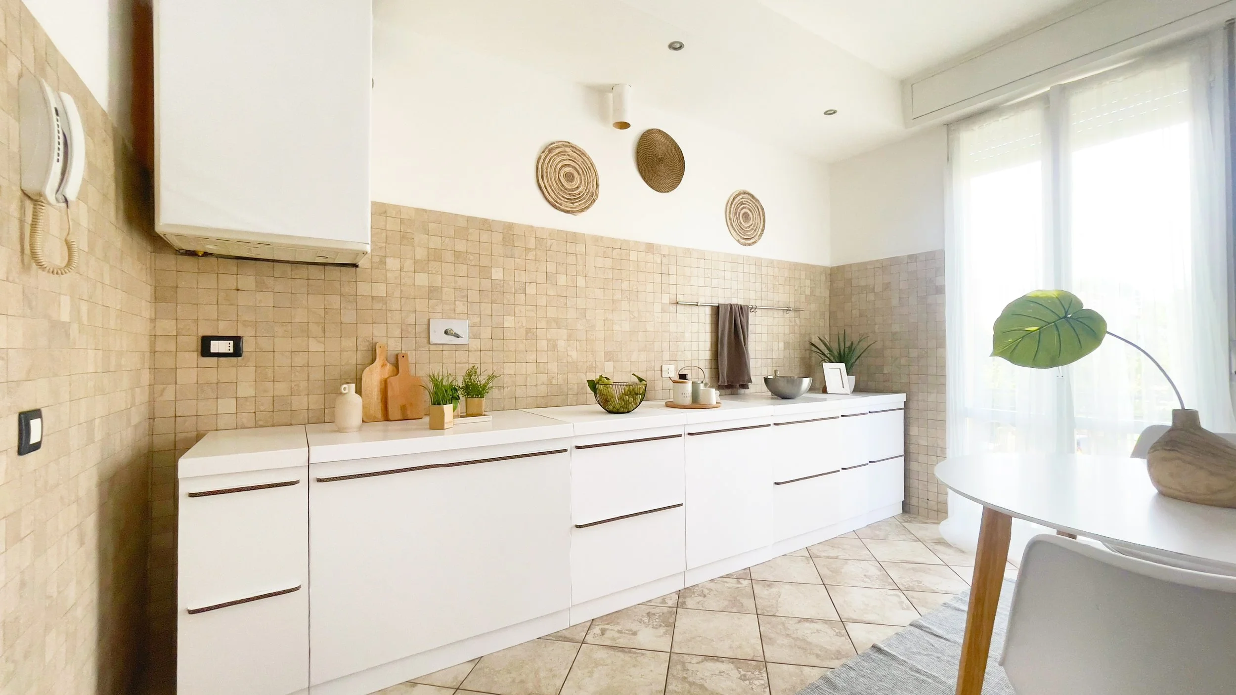 Cucina moderna con mobili bianchi, piastrelle beige, piante e decorazioni naturali.