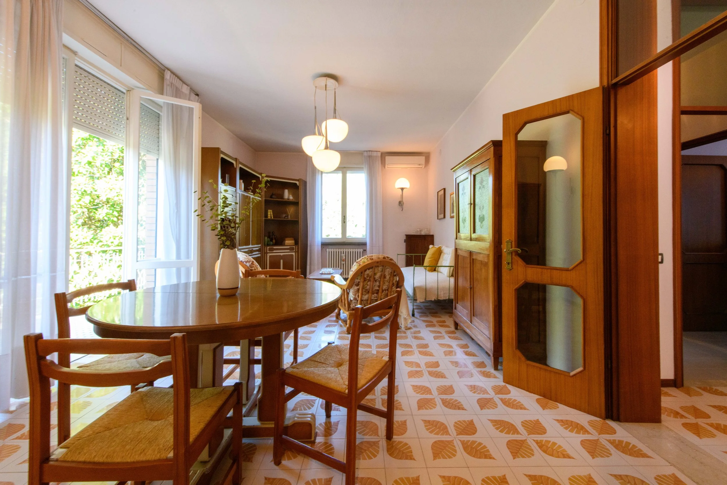 Soggiorno arredato con tavolo rotondo, sedie di legno, libreria, divano e grandi finestre con tende leggere. Stile classico con pavimento in piastrelle decorative.