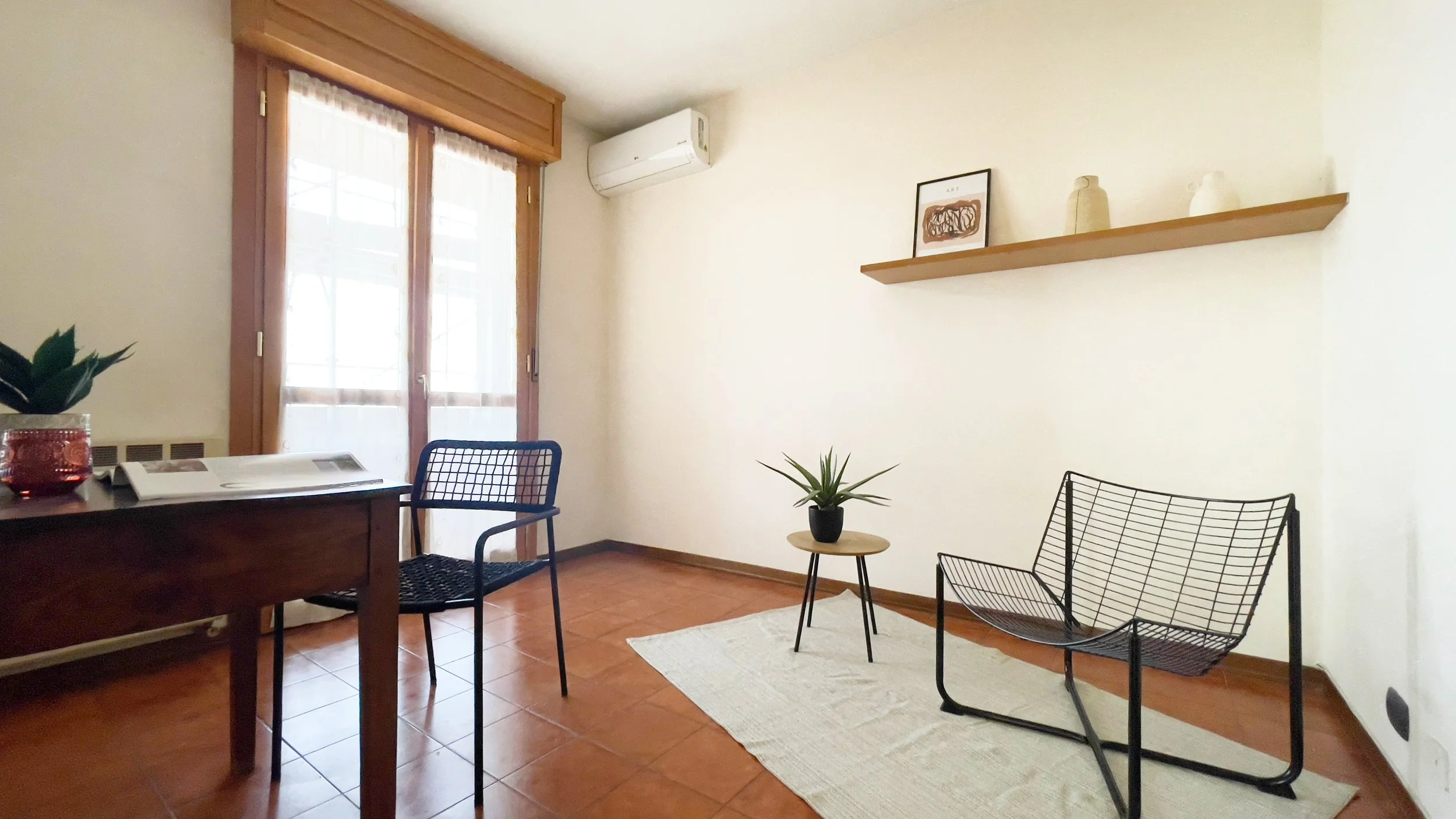 Interno di una stanza con mobili minimalisti, include una scrivania in legno, sedie in metallo, una pianta su un tavolino rotondo, una mensola con vasi e un'unità di condizionamento dell'aria. La stanza ha pavimento in piastrelle e una porta finestra