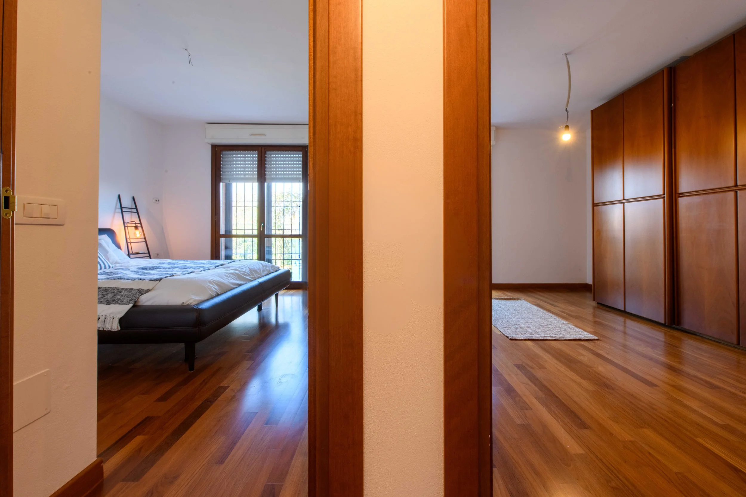 Interno di camera da letto con pavimento in legno, letto matrimoniale vicino a finestra, lampada a scala, e armadio a muro.