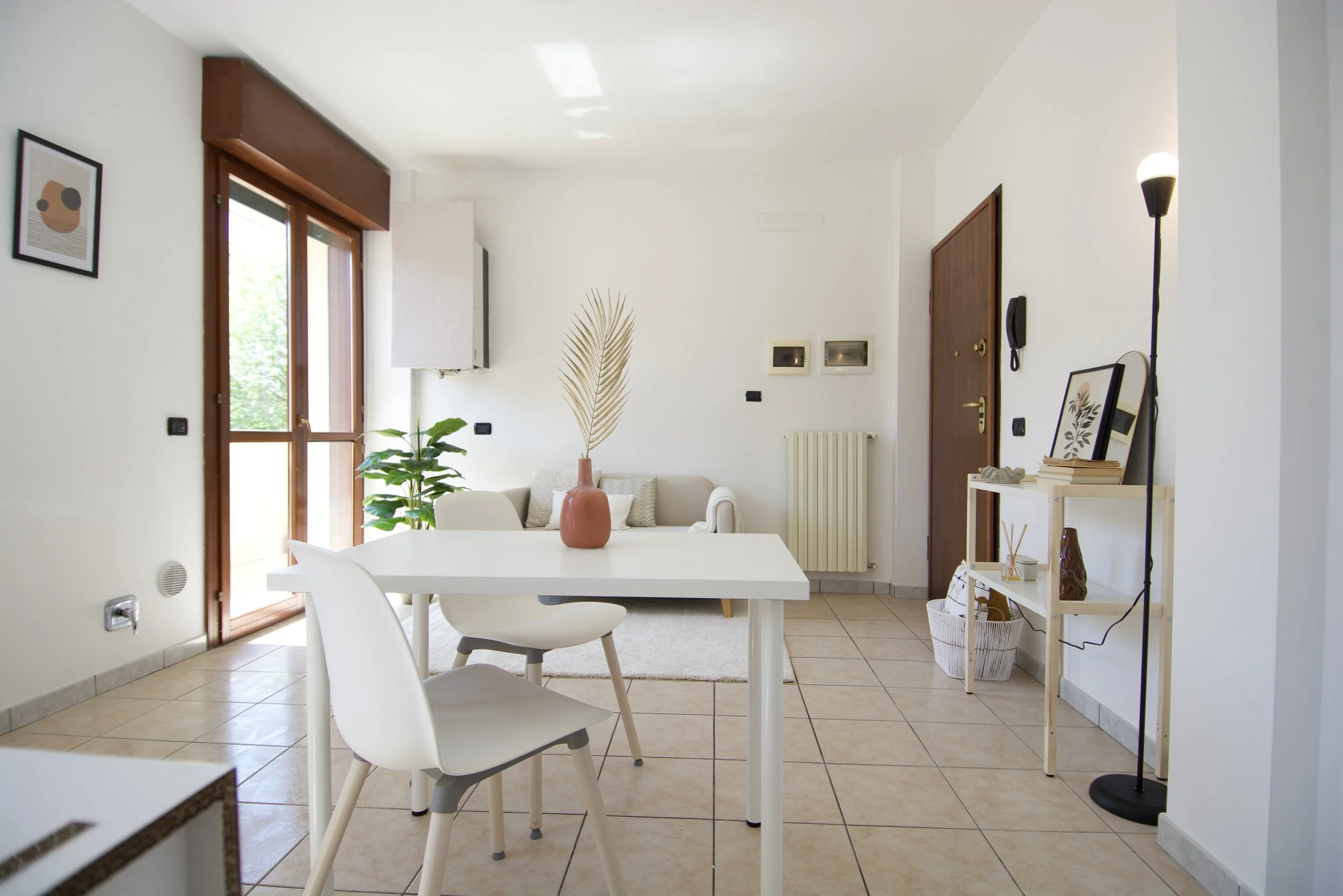 Interno di una casa moderna con arredo minimalista, tavolo bianco, sedie bianche, divano beige, pianta verde, e finestra grande con molta luce naturale.