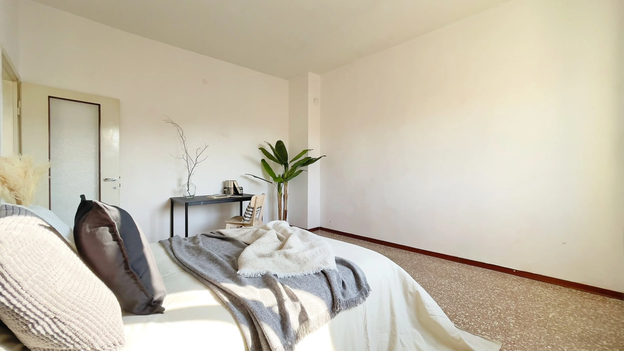 Camera da letto minimalista con letto, cuscini, coperta, scrivania con sedia, pianta e decorazioni.