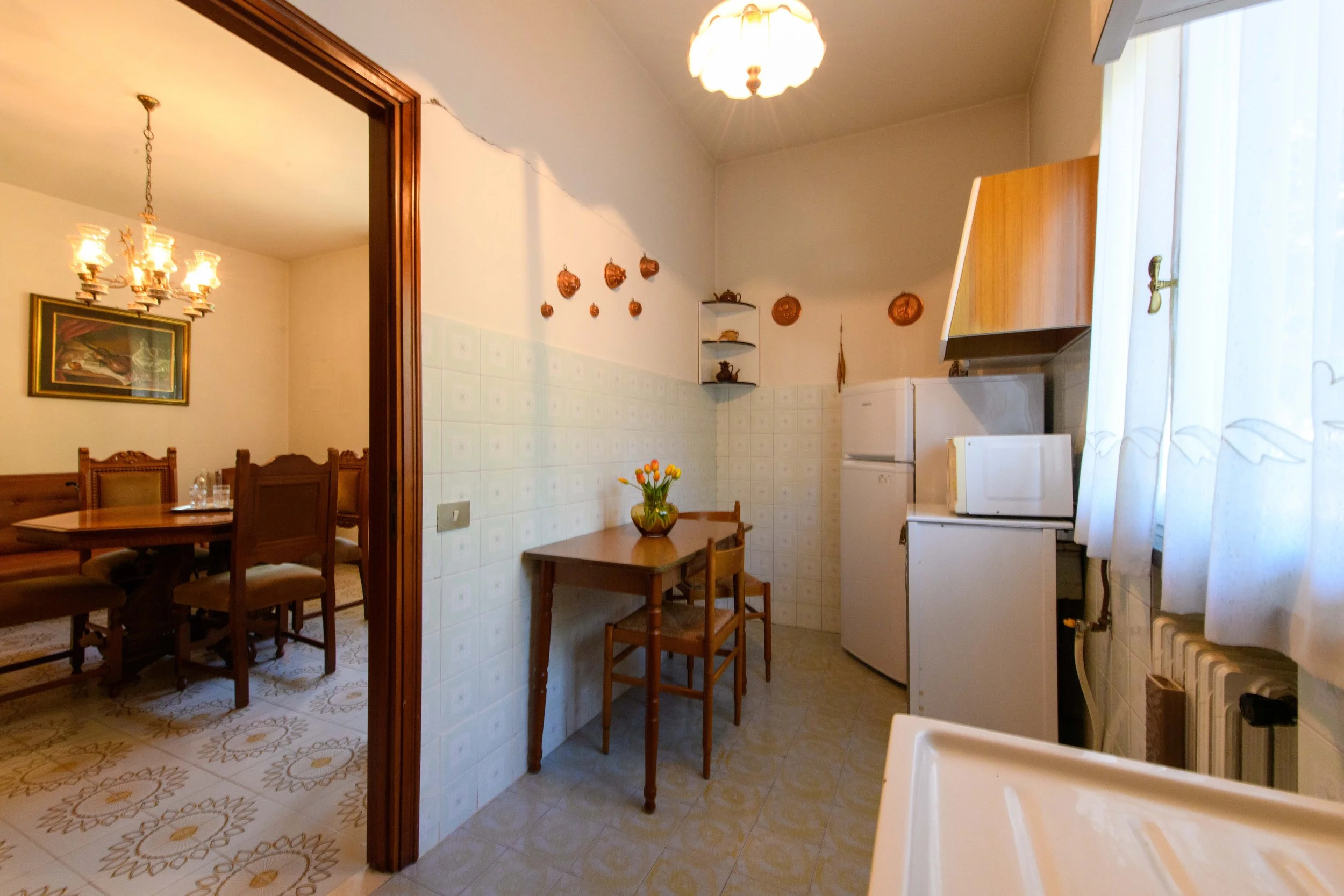 Interno di una cucina e sala da pranzo retrò con mobili in legno, tavolo, sedie e decorazioni in rame sulla parete. La cucina ha un frigorifero, microonde e una finestra con tende. Il pavimento è rivestito con piastrelle decorative. Luce pendente sop