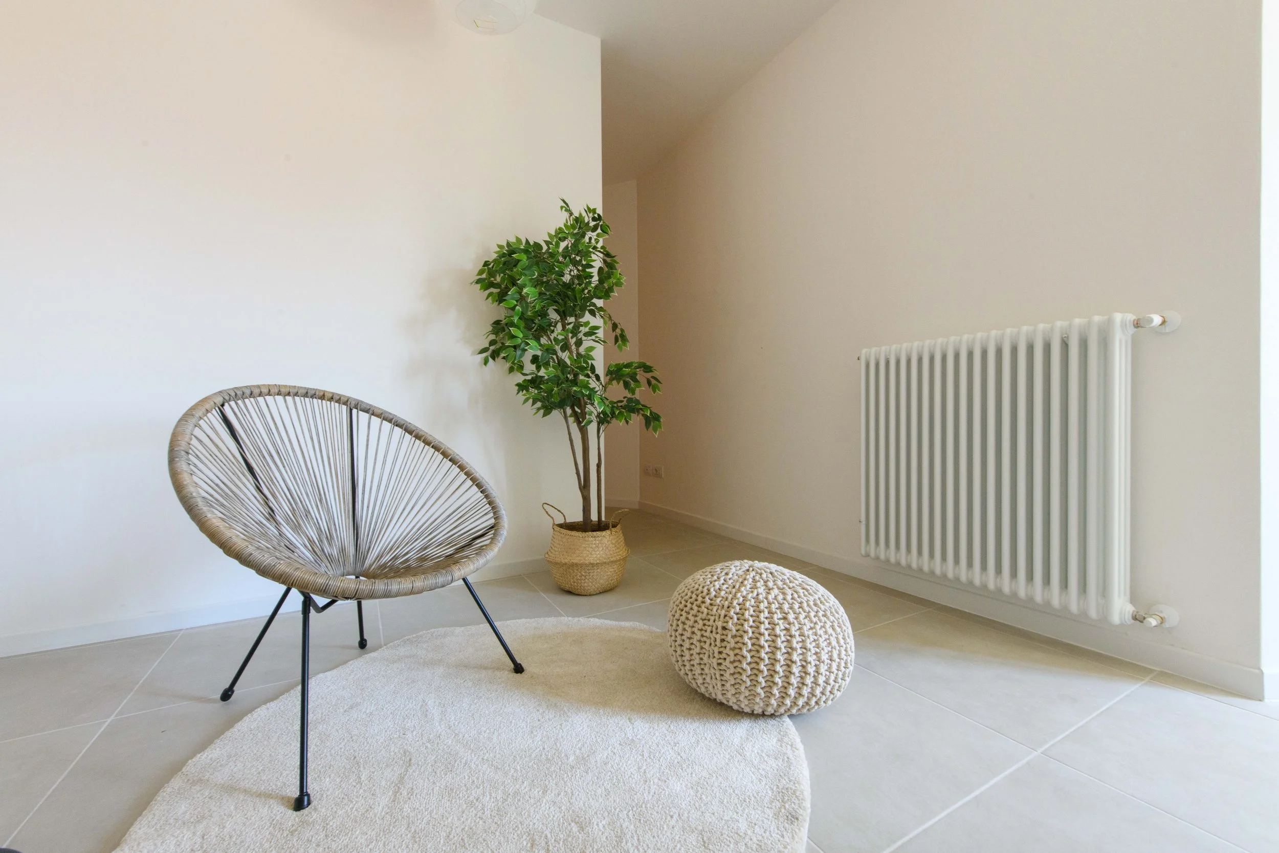 Interno di una stanza minimalista con una sedia design in metallo e corda, una pianta in un vaso intrecciato, un pouf in maglia e un tappeto chiaro su un pavimento piastrellato. Sul muro bianco c'è un radiatore moderno.