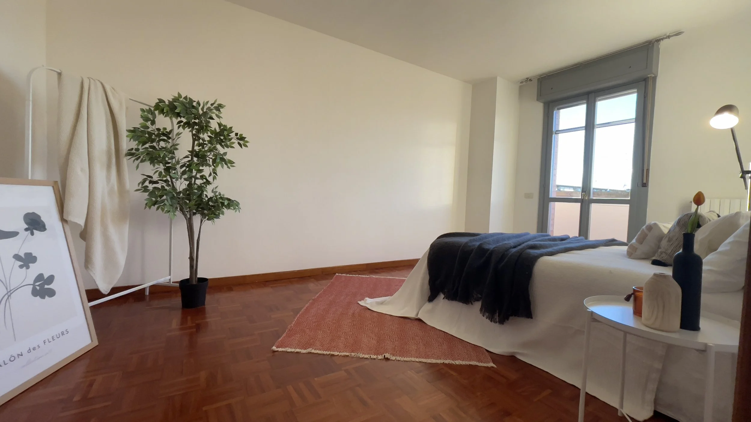 Camera da letto minimalista con parquet, letto matrimoniale con coperte bianche e nere, tavolino bianco, pianta in vaso, finestra, tappeto rosa e quadro appoggiato al muro.
