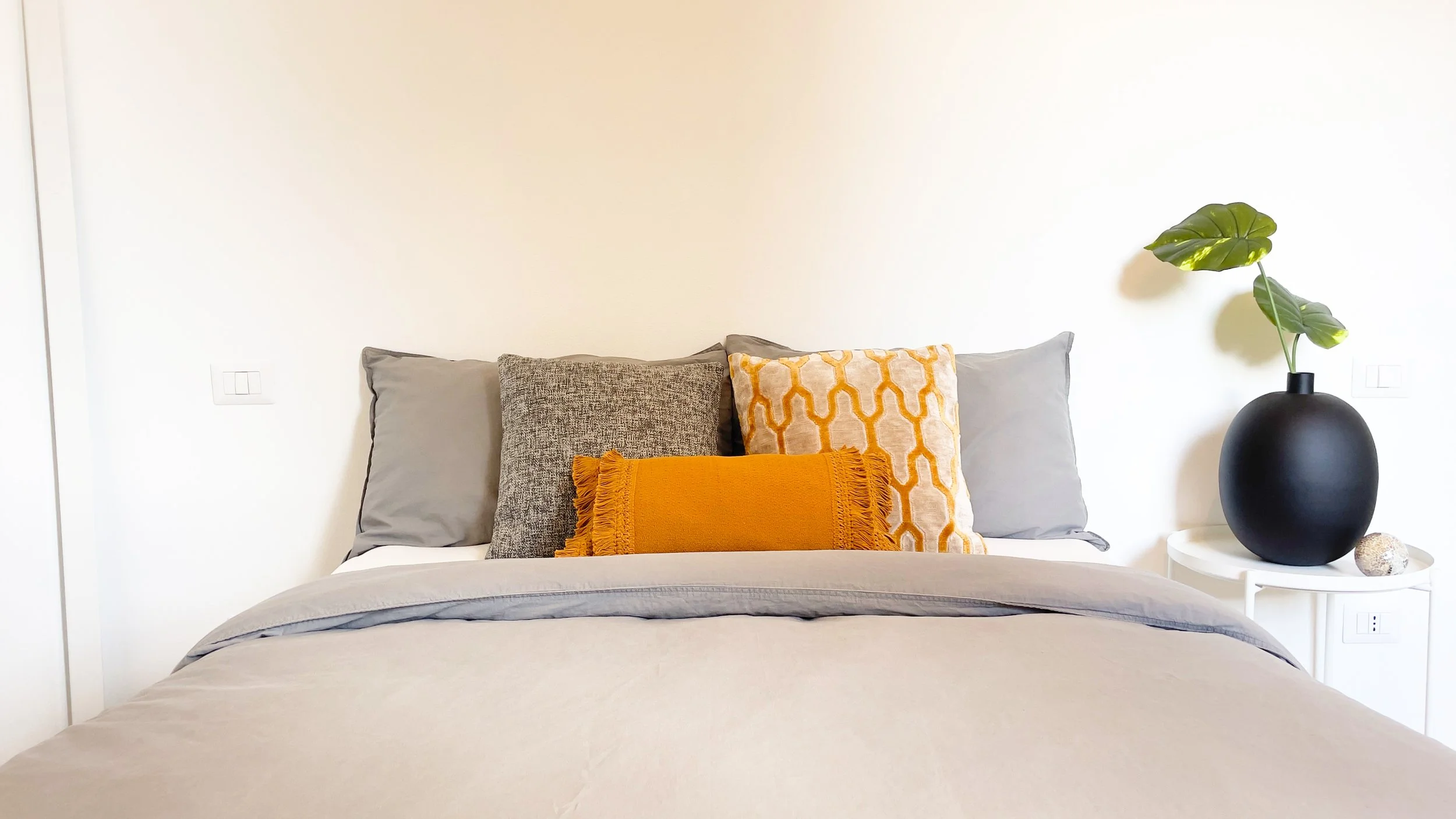 Camera da letto moderna con letto matrimoniale e cuscini multicolore, un vaso nero con pianta verde su un comodino bianco.