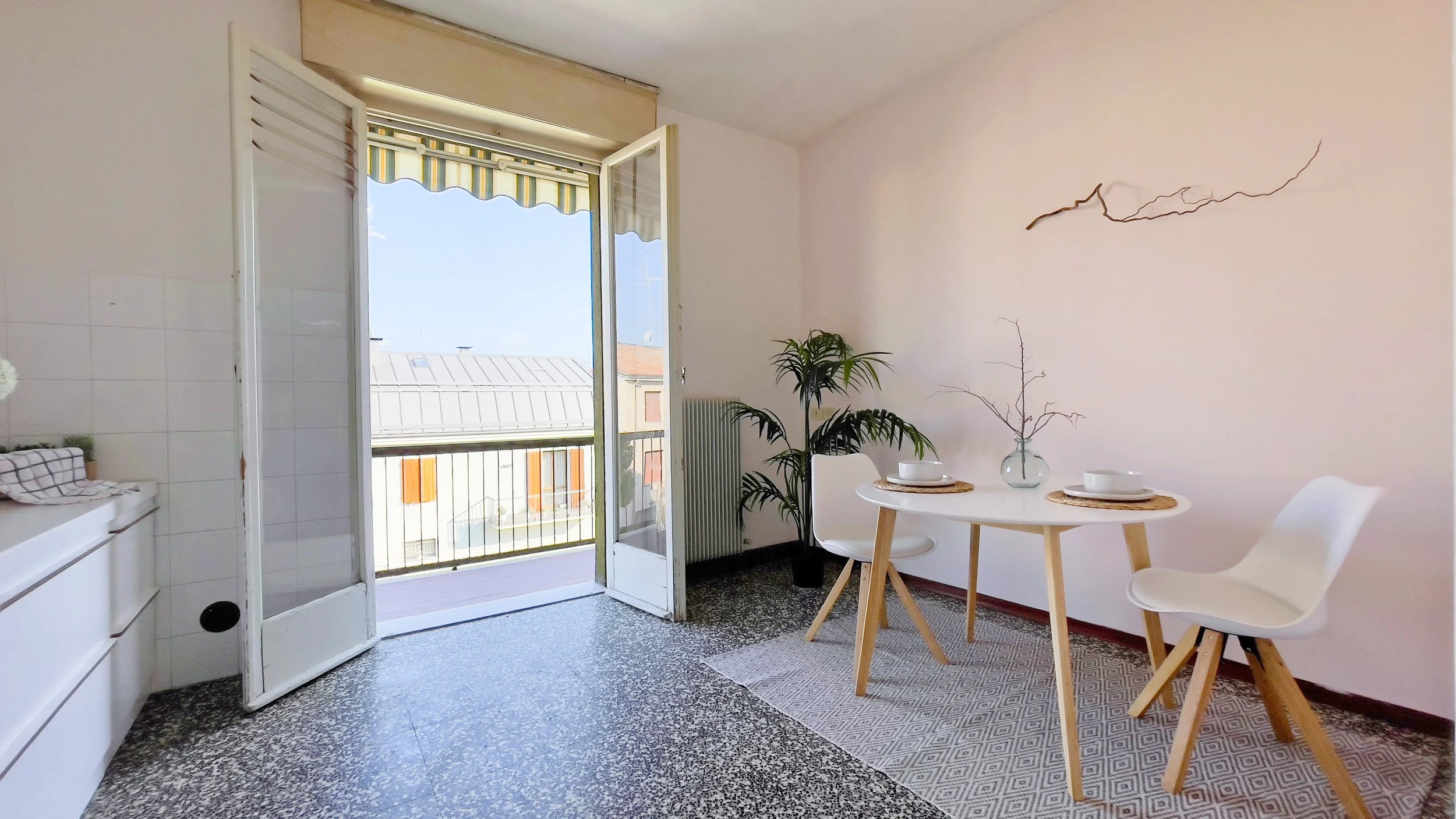 Interno di una stanza luminosa con tavolo, sedie bianche, pianta decorativa, finestra aperta su balcone e muro con decorazione.
