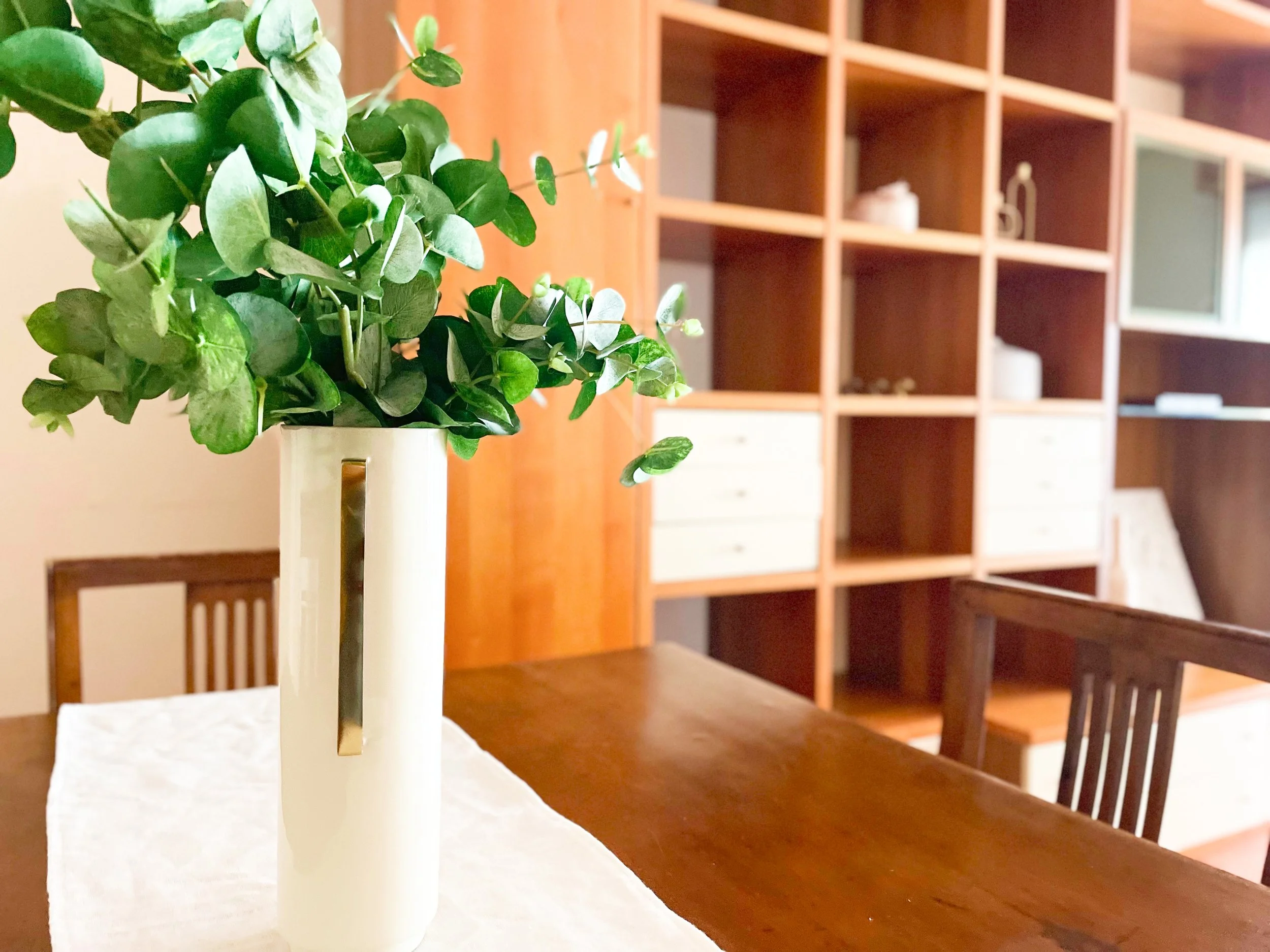 Vaso bianco con foglie verdi su un tavolo di legno davanti a una libreria con scaffali aperti.