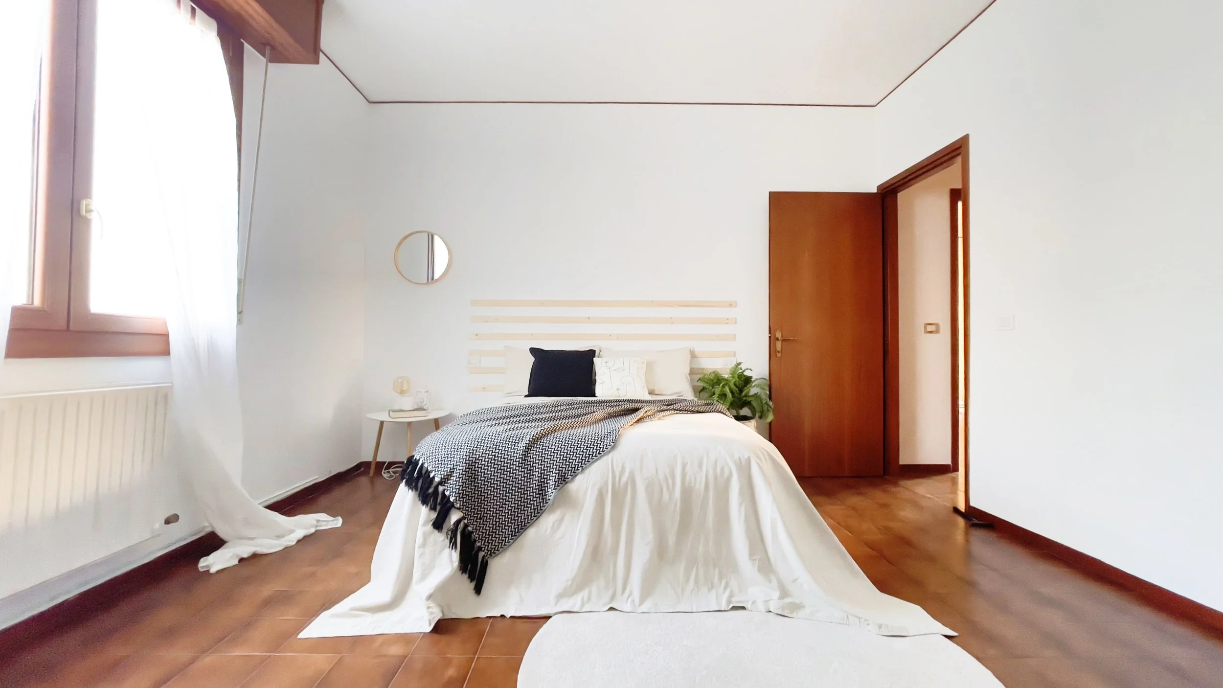 Camera da letto minimalista con letto matrimoniale, copriletto bianco, cuscini neri, un comodino con lampada, pianta e finestre con tende bianche.