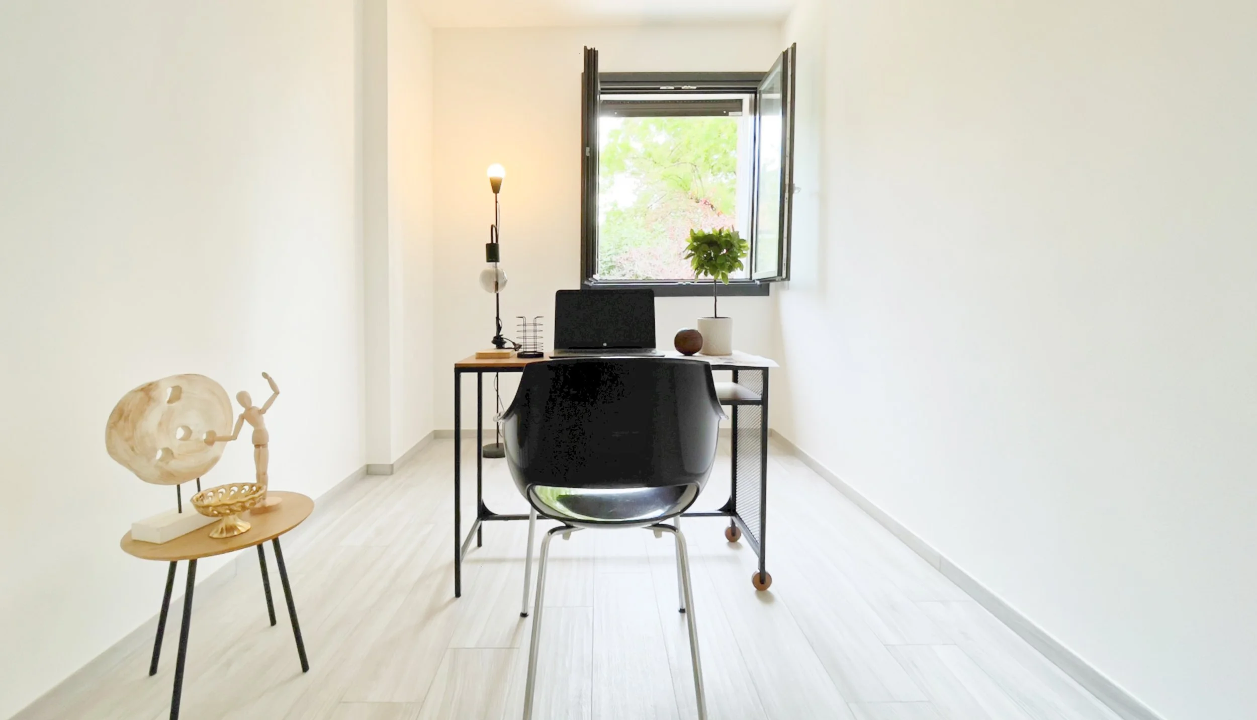Ufficio minimalista con una scrivania moderna, sedia nera, laptop, pianta vasetto, lampada da tavolo, finestra aperta e decorazioni moderne su un tavolino laterale.