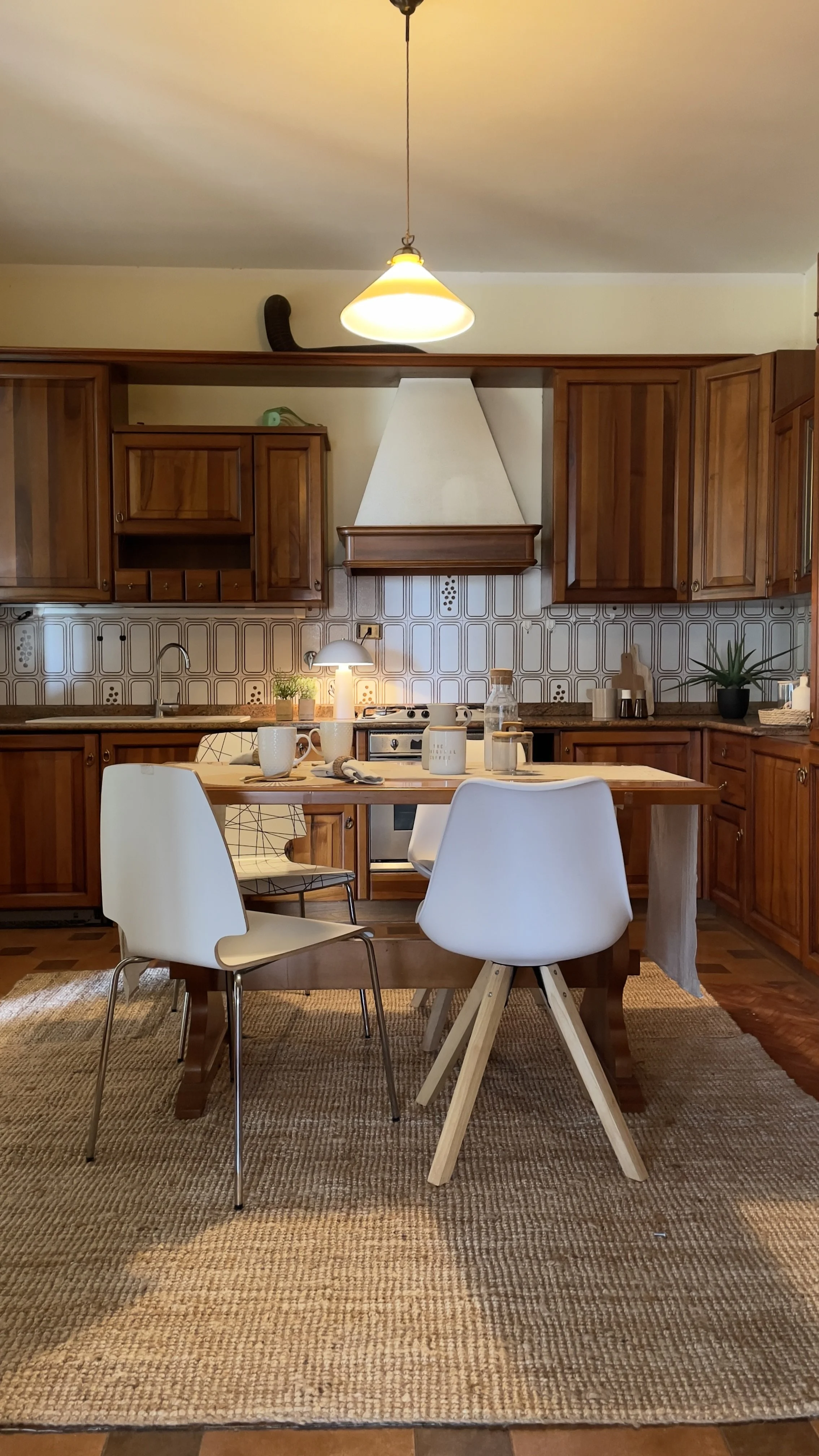 Cucina con mobili in legno, tavolo apparecchiato, sedie bianche e lampadario a sospensione.