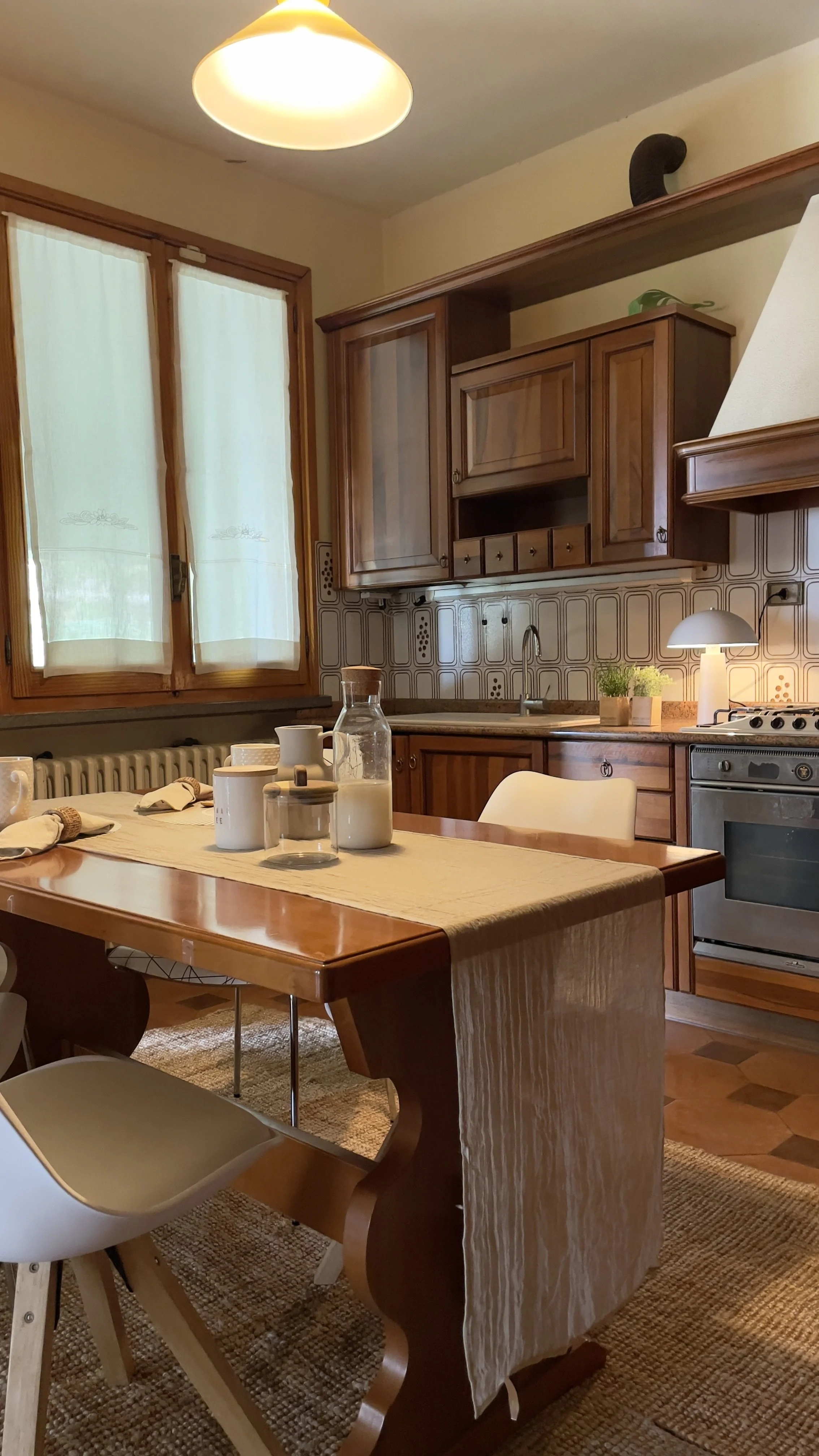 Cucina rustica con mobili in legno, tavolo apparecchiato con tovaglia, bottiglia di latte, tazze, e sedie moderne. Finestra con tende bianche e forno a gas visibile.