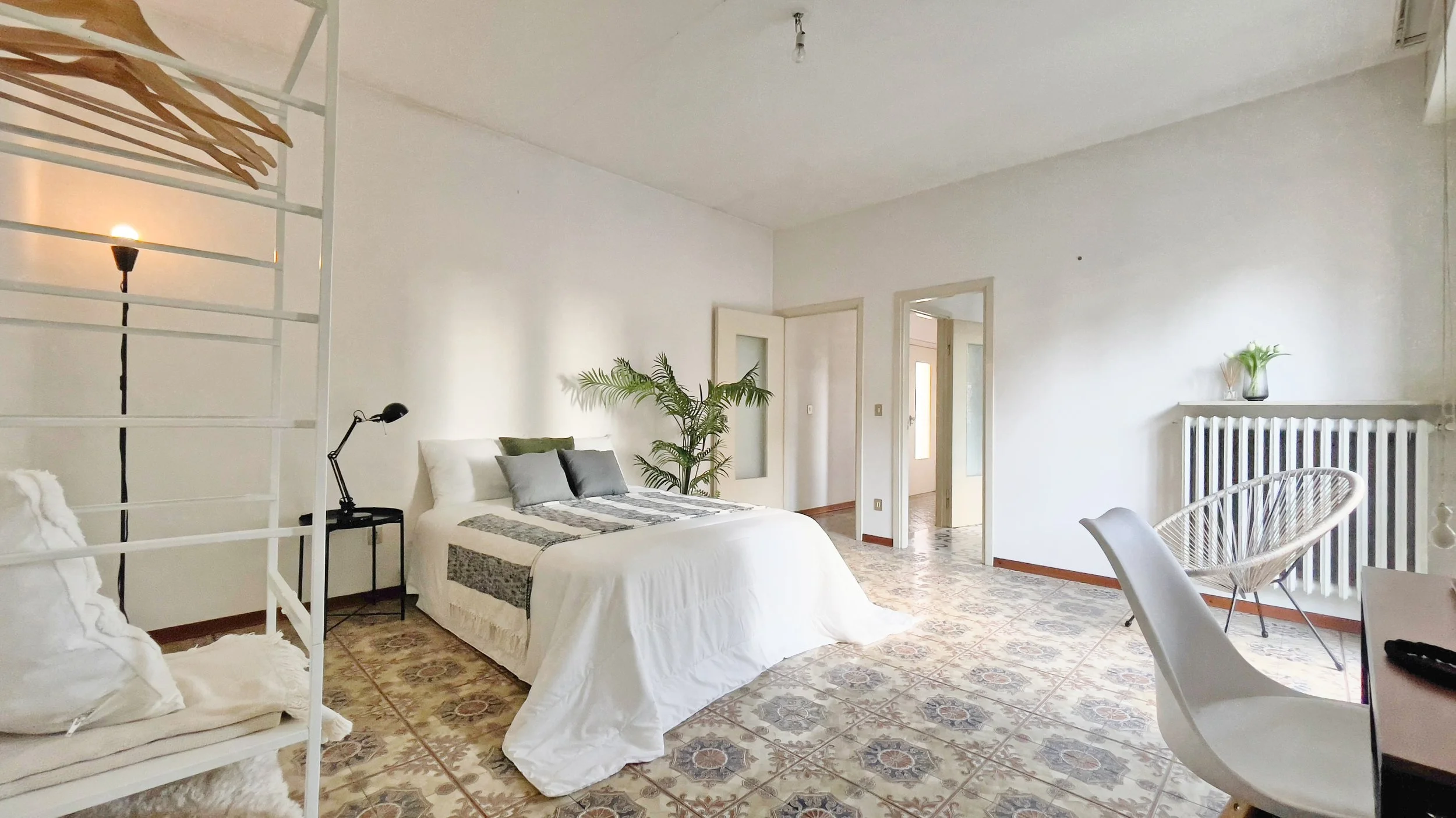 Camera da letto luminosa con letto matrimoniale, lampada da terra nera, pianta decorativa, sedia in stile moderno e pavimento piastrellato.