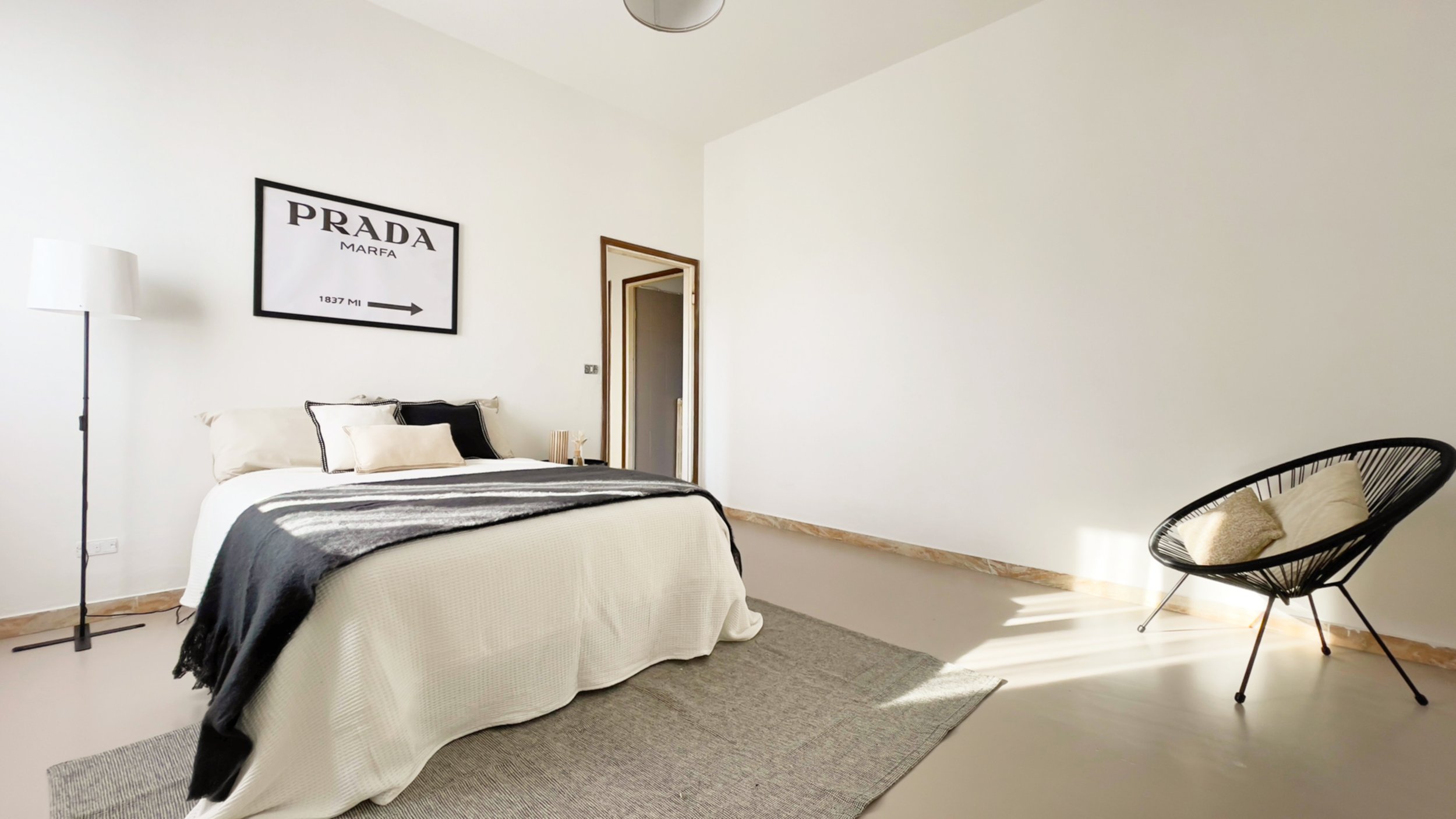 Camera da letto minimalista con letto matrimoniale, quadro su parete con scritta "Prada Marfa", lampada a piantana, sedia moderna con cuscino, arredamento in toni neutri e luminosi.