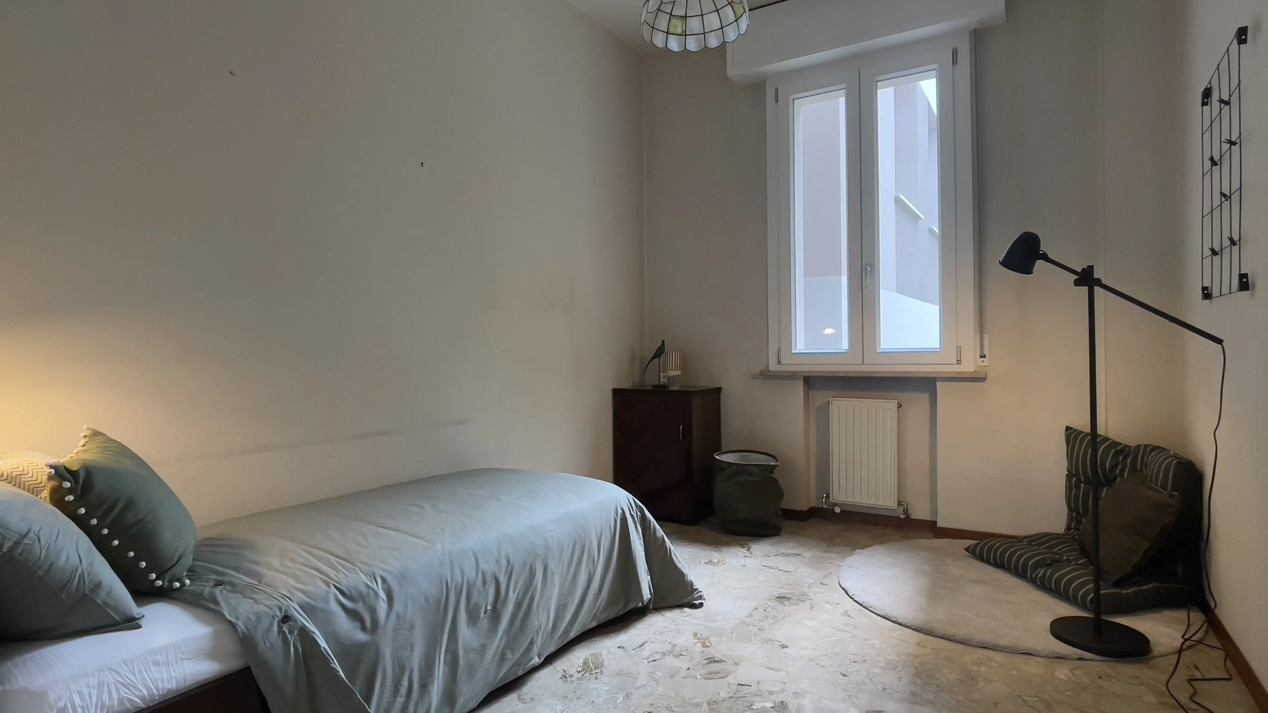 Camera da letto minimalista con letto singolo, cuscini verdi, comodino, finestra, tappetino rotondo e lampada da terra.