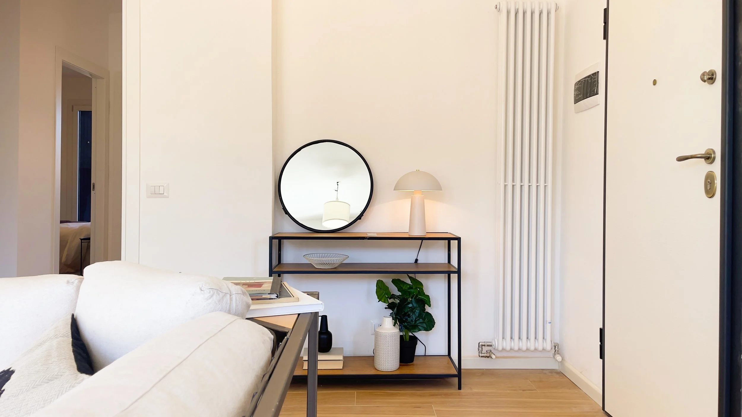 Angolo di un soggiorno moderno con un divano bianco, specchio rotondo sopra una mensola, lampada da tavolo bianca, e pianta decorativa. Sul pavimento parquet chiaro.