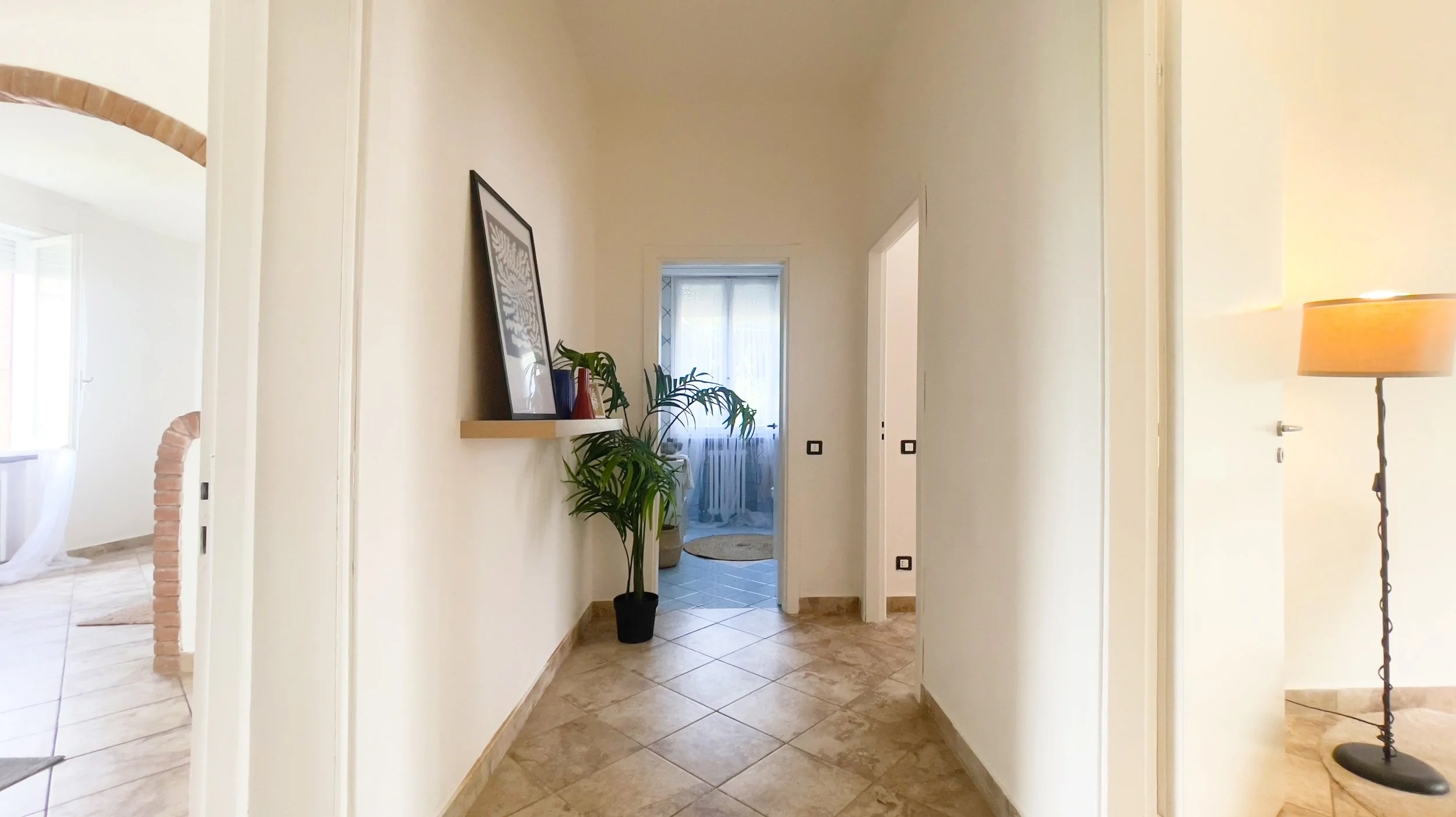 Corridoio di un appartamento con pavimento piastrellato, lampada da terra a destra, piante in vaso e quadro su mensola a sinistra, e porta in fondo che conduce a una stanza con finestre.