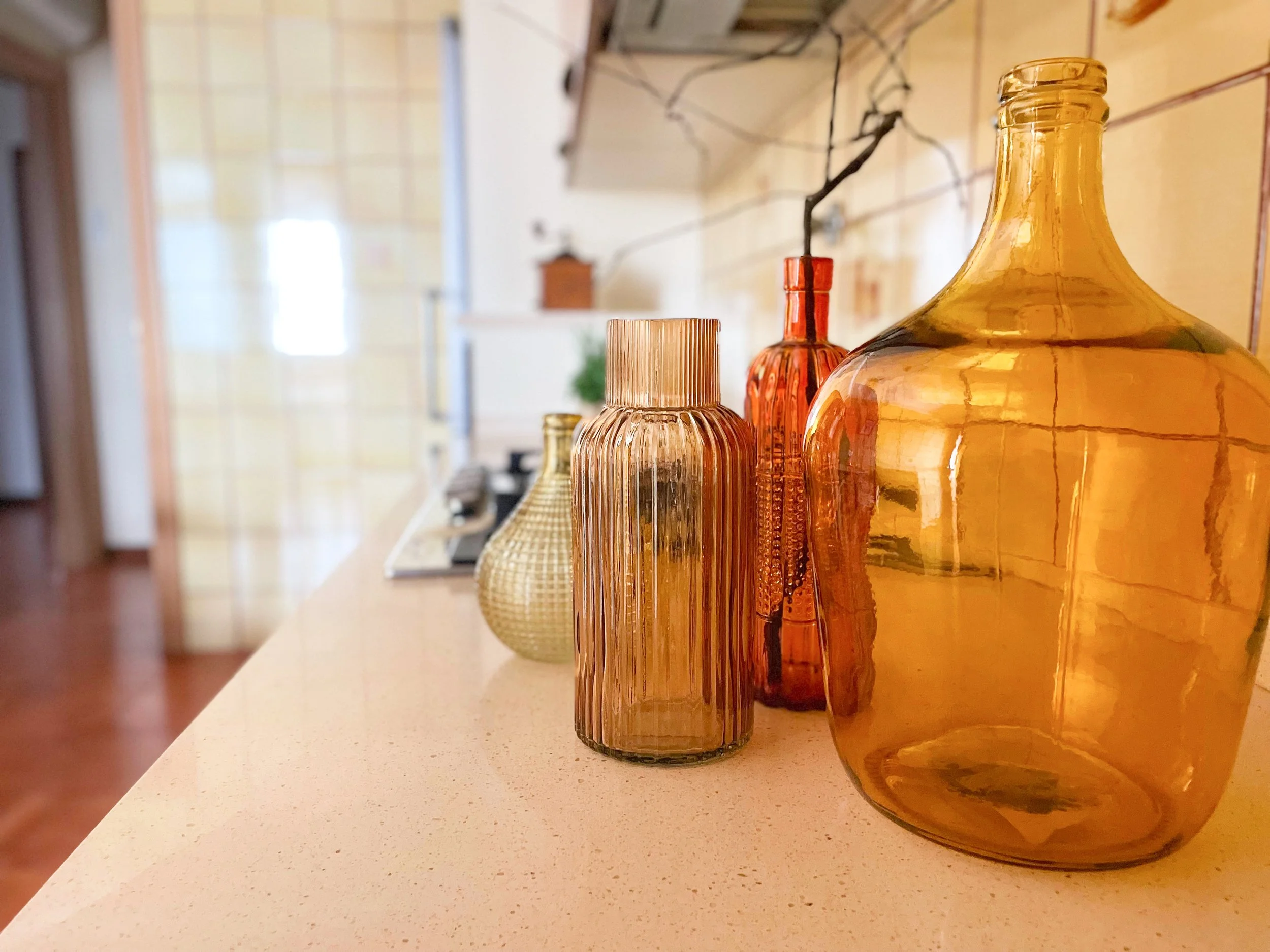 Bottiglie di vetro di vari colori e forme su un ripiano, sfondo di una cucina con piastrelle gialle.
