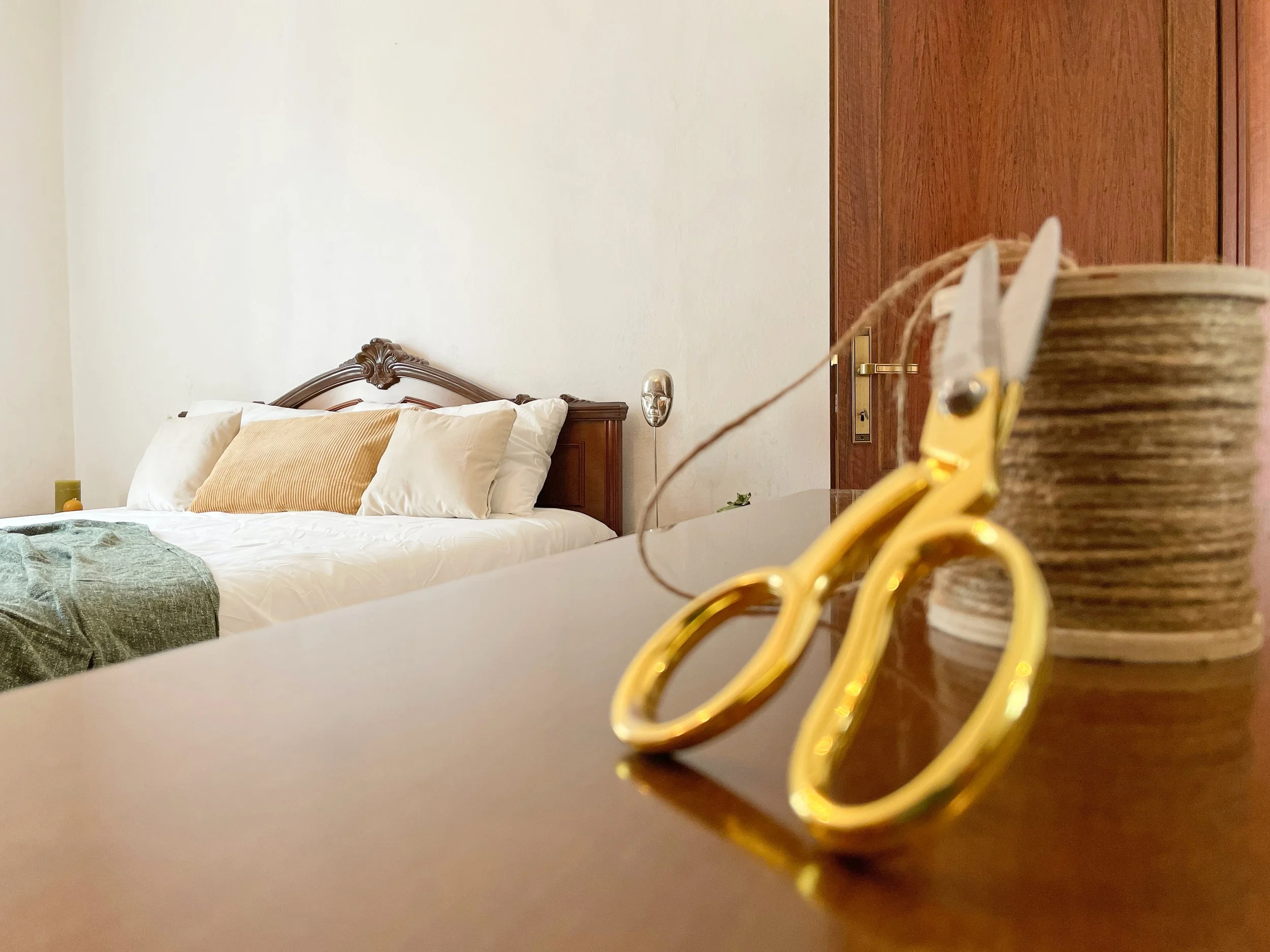 Interno di una camera da letto con un letto matrimoniale coperto da cuscini bianchi e un copriletto verde. In primo piano, un paio di forbici dorate e un rocchetto di filo su un tavolo di legno.
