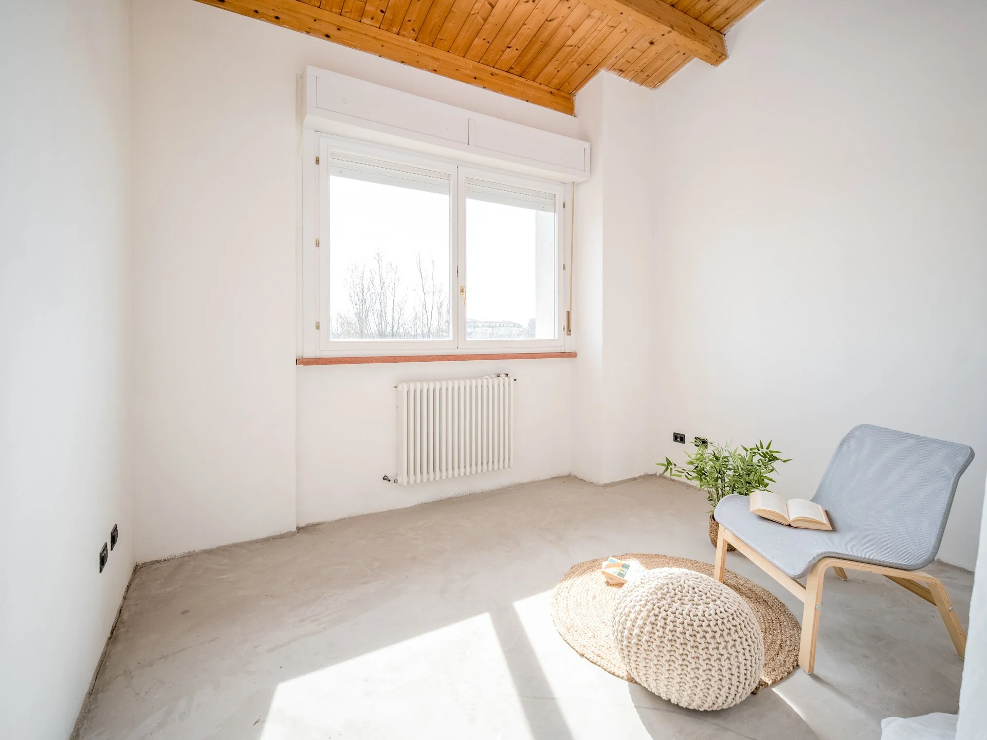 Stanza minimalista con pareti bianche, soffitto in legno, finestra grande, sedia moderna grigia, tappeto rotondo in paglia, cuscino lavorato a maglia e pianta verde decorativa.