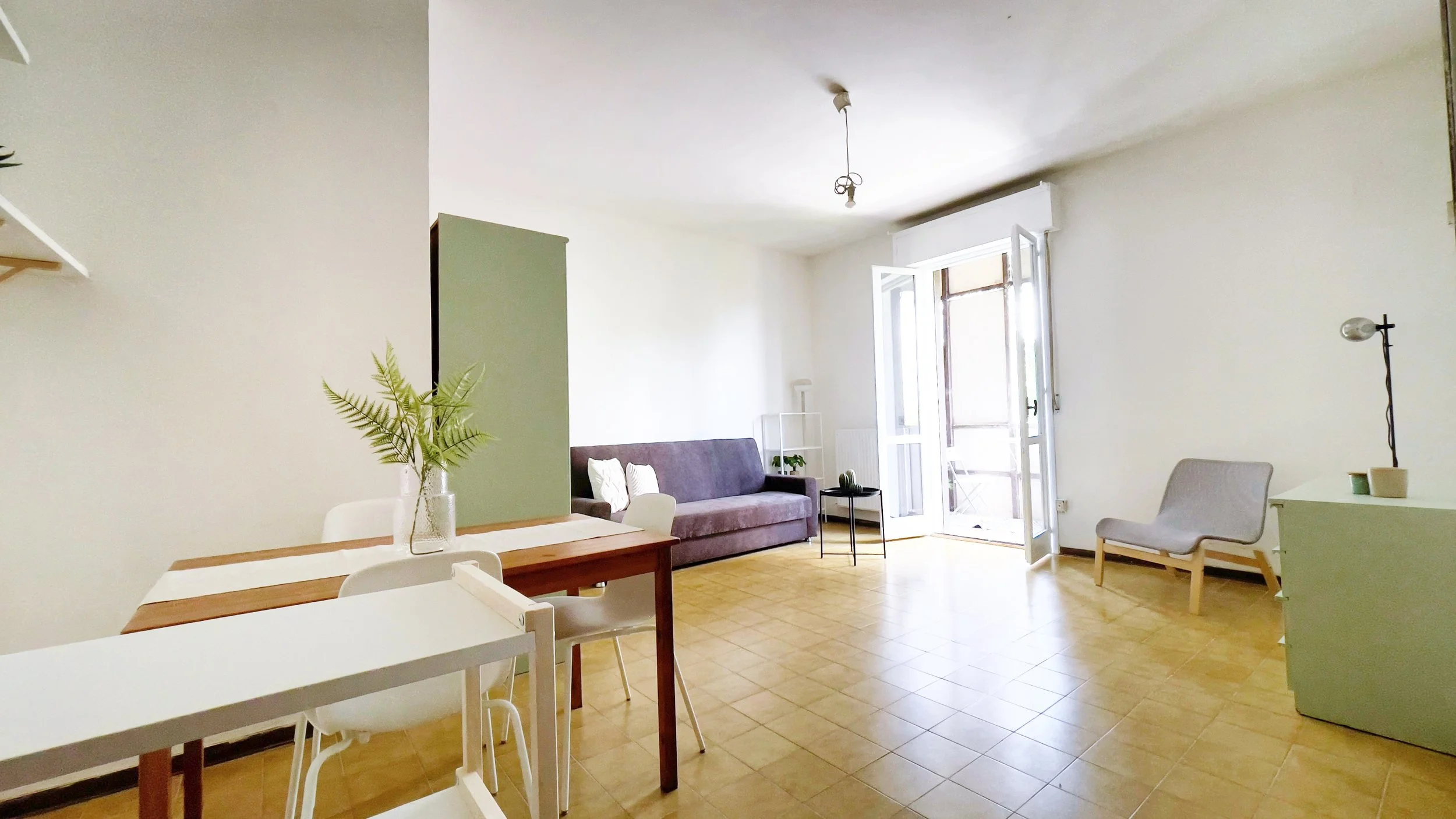 Soggiorno moderno e luminoso con tavolo da pranzo in legno, sedie bianche, divano grigio, poltrona, mobile verde e pianta decorativa. Porta-finestra aperta e pavimento in piastrelle beige chiaro.