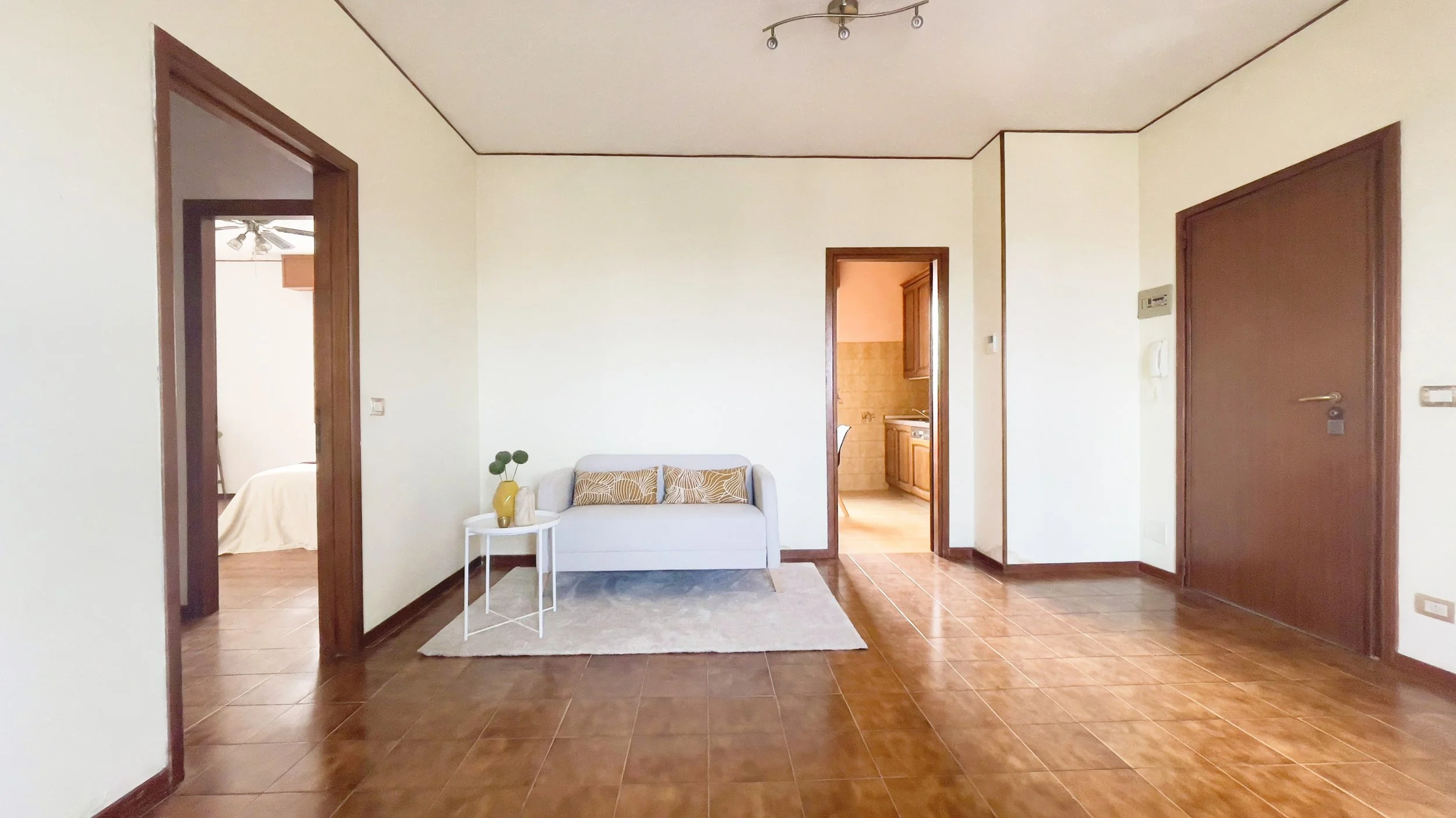 Soggiorno accogliente con divano, tavolino, tappeto e due porte che conducono a camere adiacenti. Pavimento in legno e semplice illuminazione a soffitto.