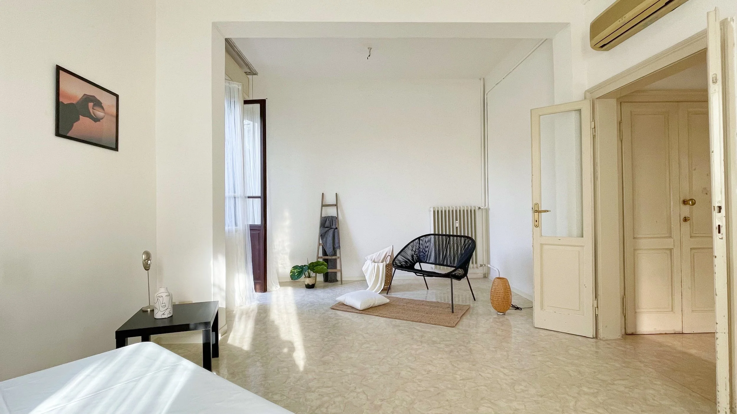 Stanza luminosa con arredamento minimalista, una sedia nera, un tavolino con lampada e decorazioni, una scala di legno come portaoggetti, una pianta e un cuscino bianco sul pavimento.