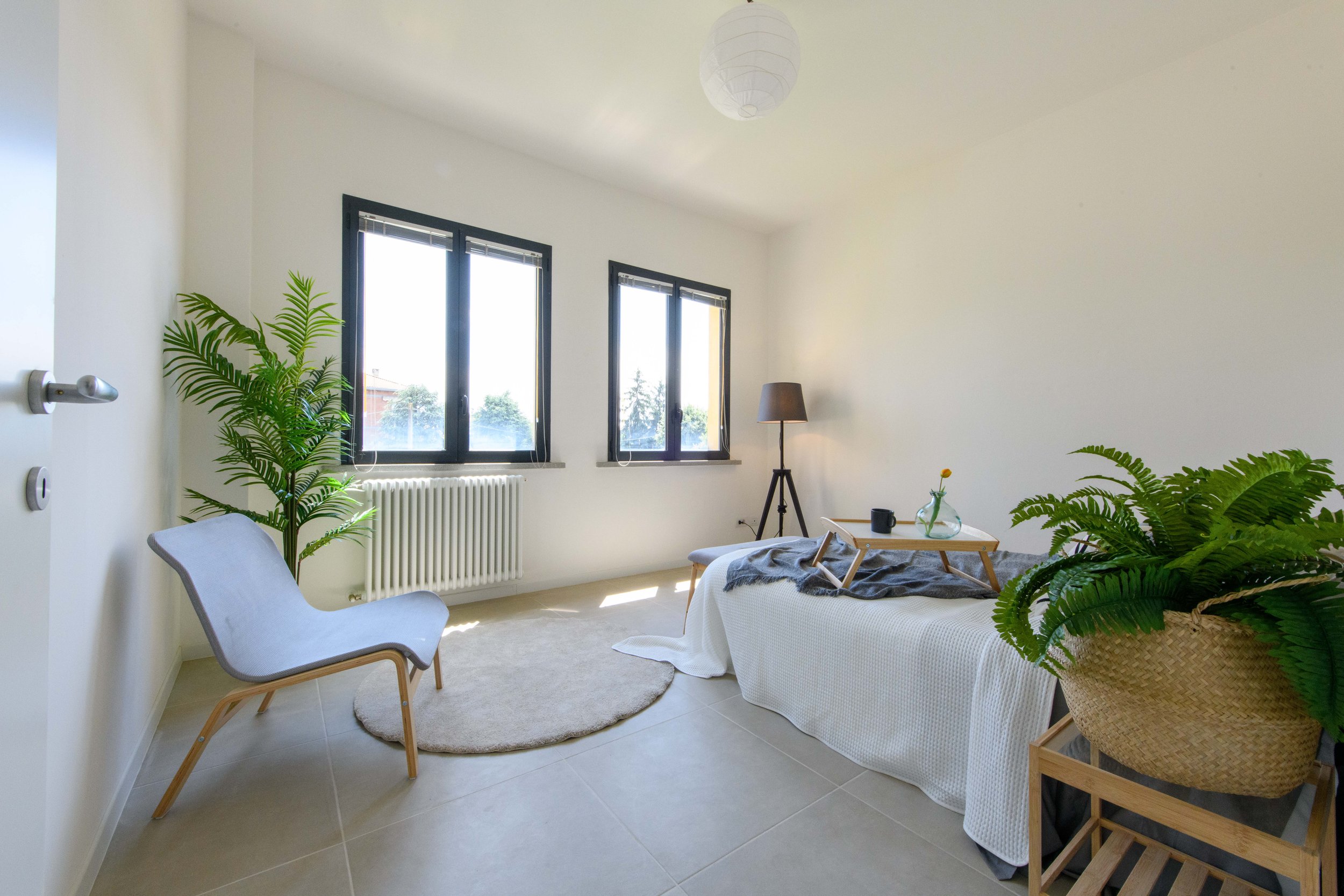 Camera da letto moderna e minimalista con una chaise longue, una pianta in vaso, un letto singolo coperto da un copriletto bianco, un tavolino sul letto, un tappeto rotondo beige, una lampada da terra e finestre luminose.