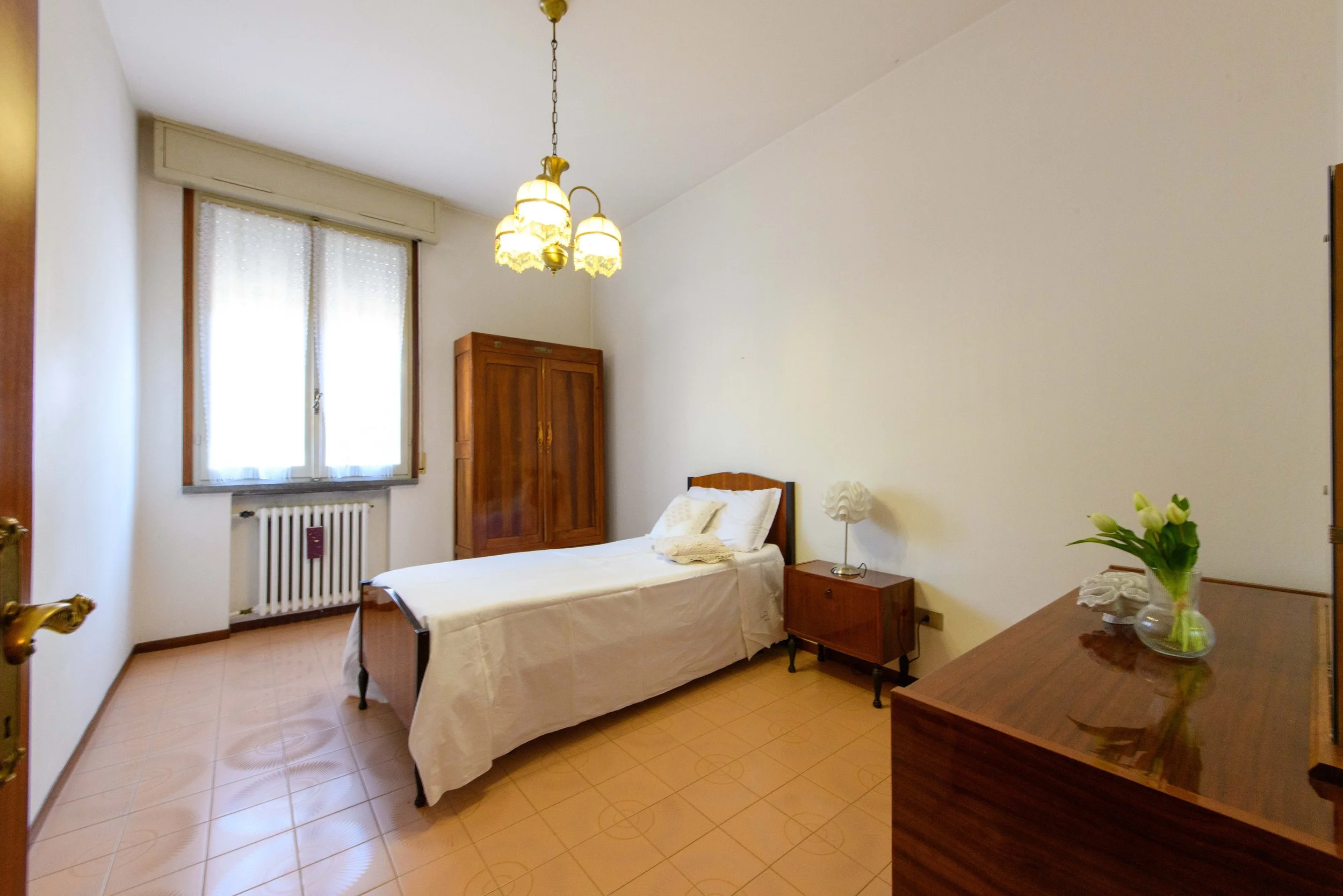 Camera da letto con letto singolo, armadio in legno, comodino con lampada, fiori in vaso, e finestra con tende.