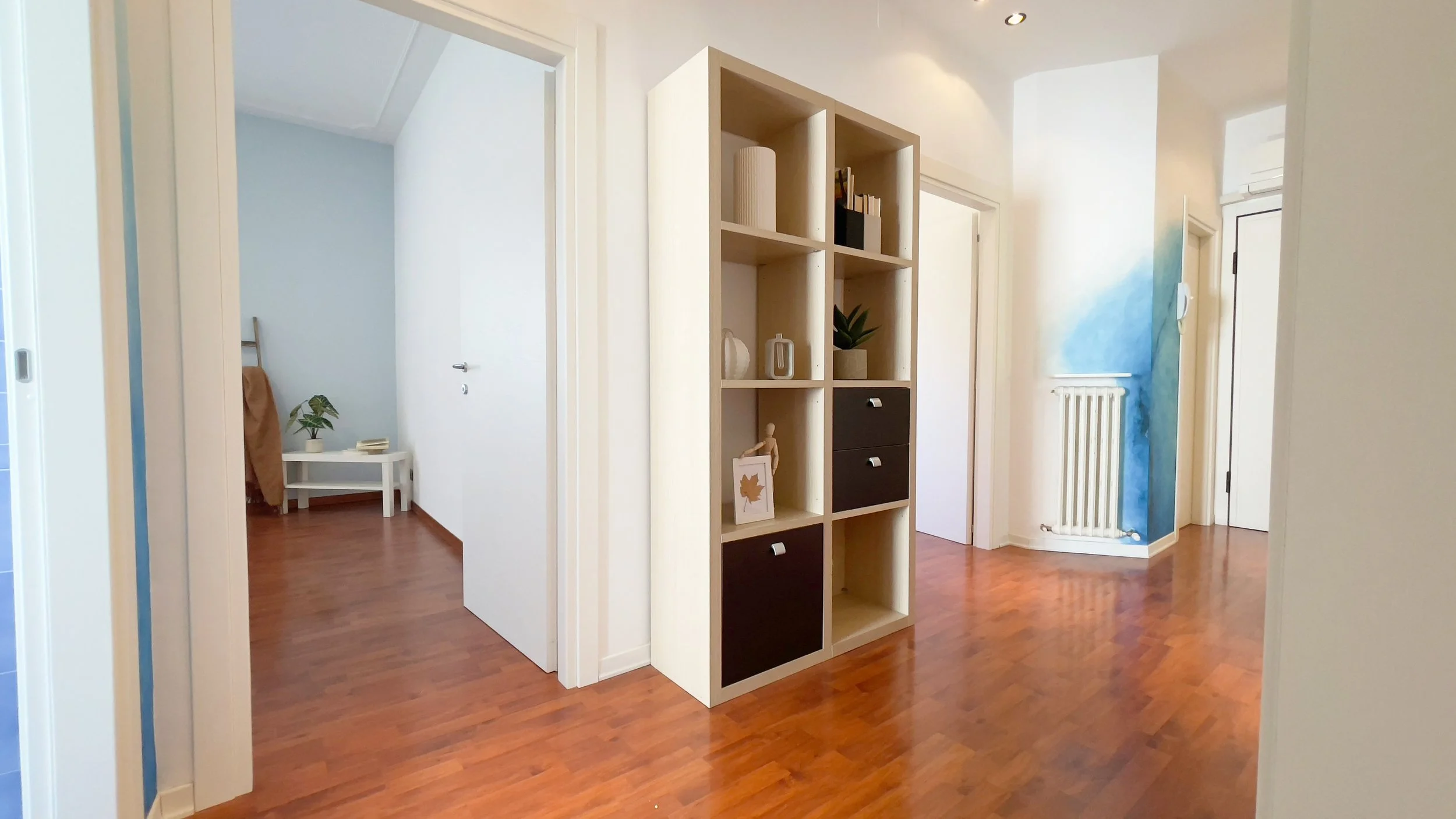 Corridoio luminoso con pavimento in legno, scaffale moderno e porte aperte che conducono a stanze adiacenti.