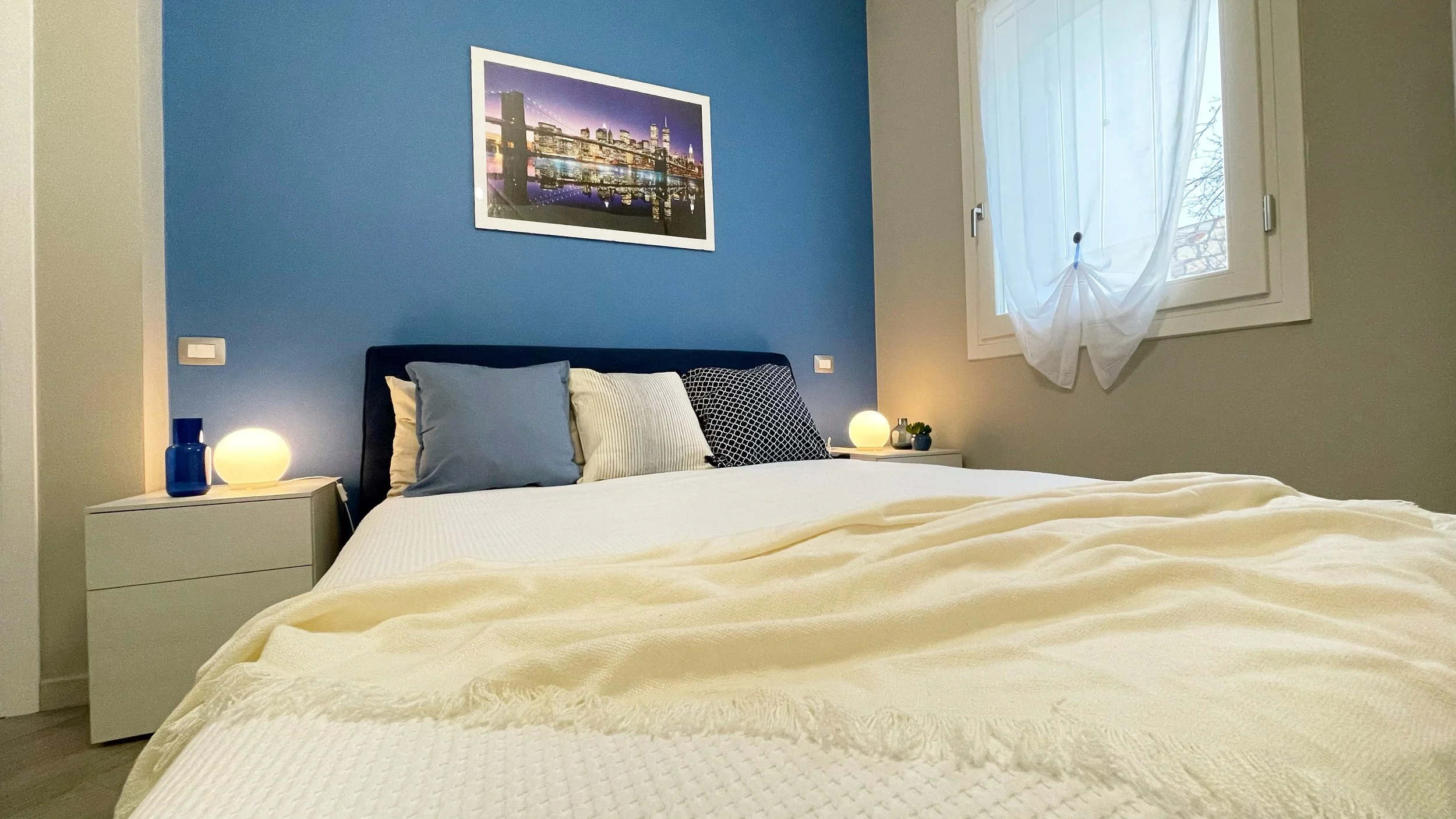 Camera da letto moderna con pareti blu, letto matrimoniale con coperte bianche, e comodini con lampade accese. Sul muro c'è un quadro di un skyline urbano.