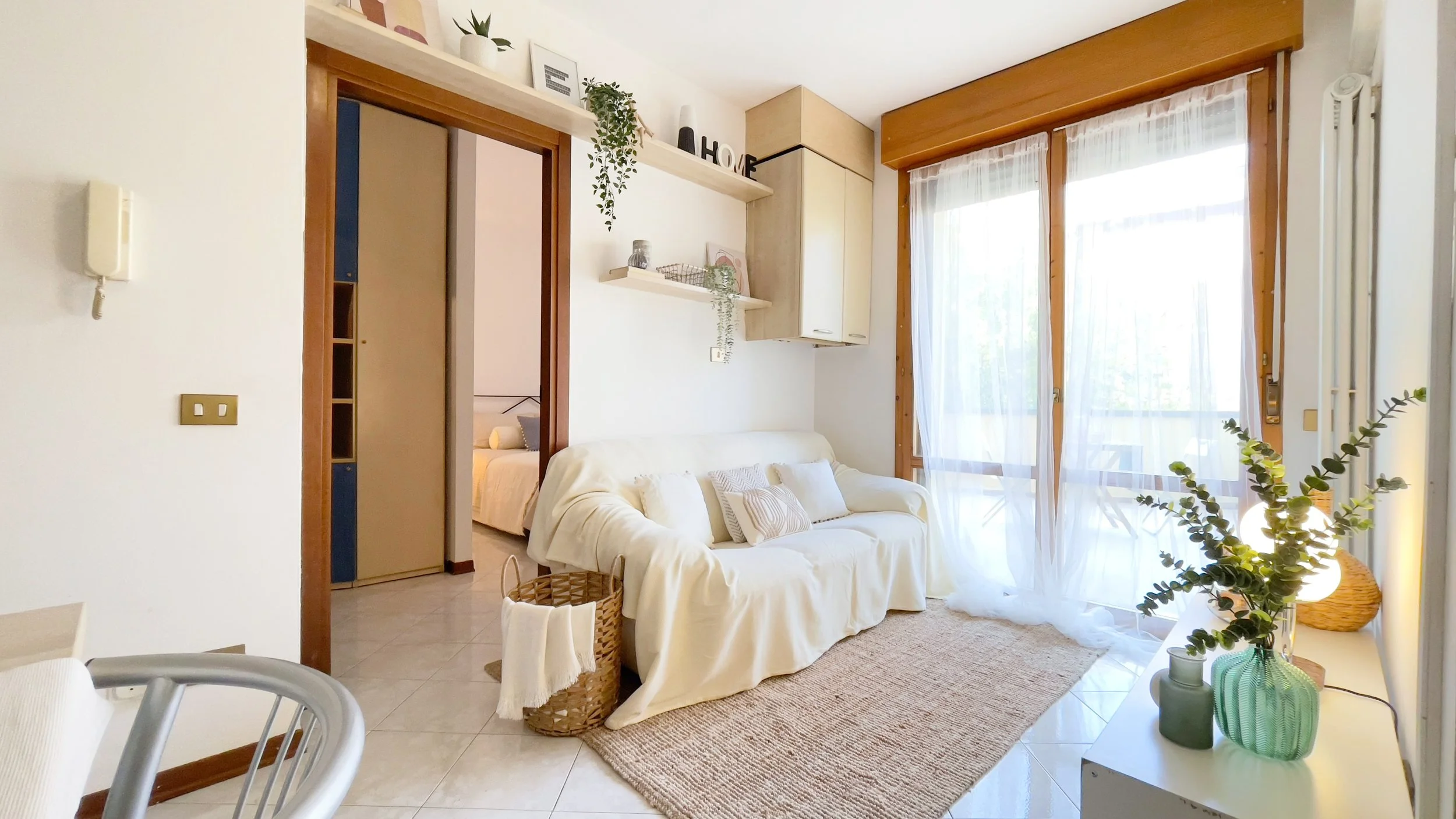 Soggiorno luminoso con divano bianco e cuscini, accanto a un grande balcone con tende trasparenti. Ci sono mensole con piante e decorazioni sopra il divano, un cesto con coperta e un vaso con rami verdi su un tavolino illuminato da una lampada sferic