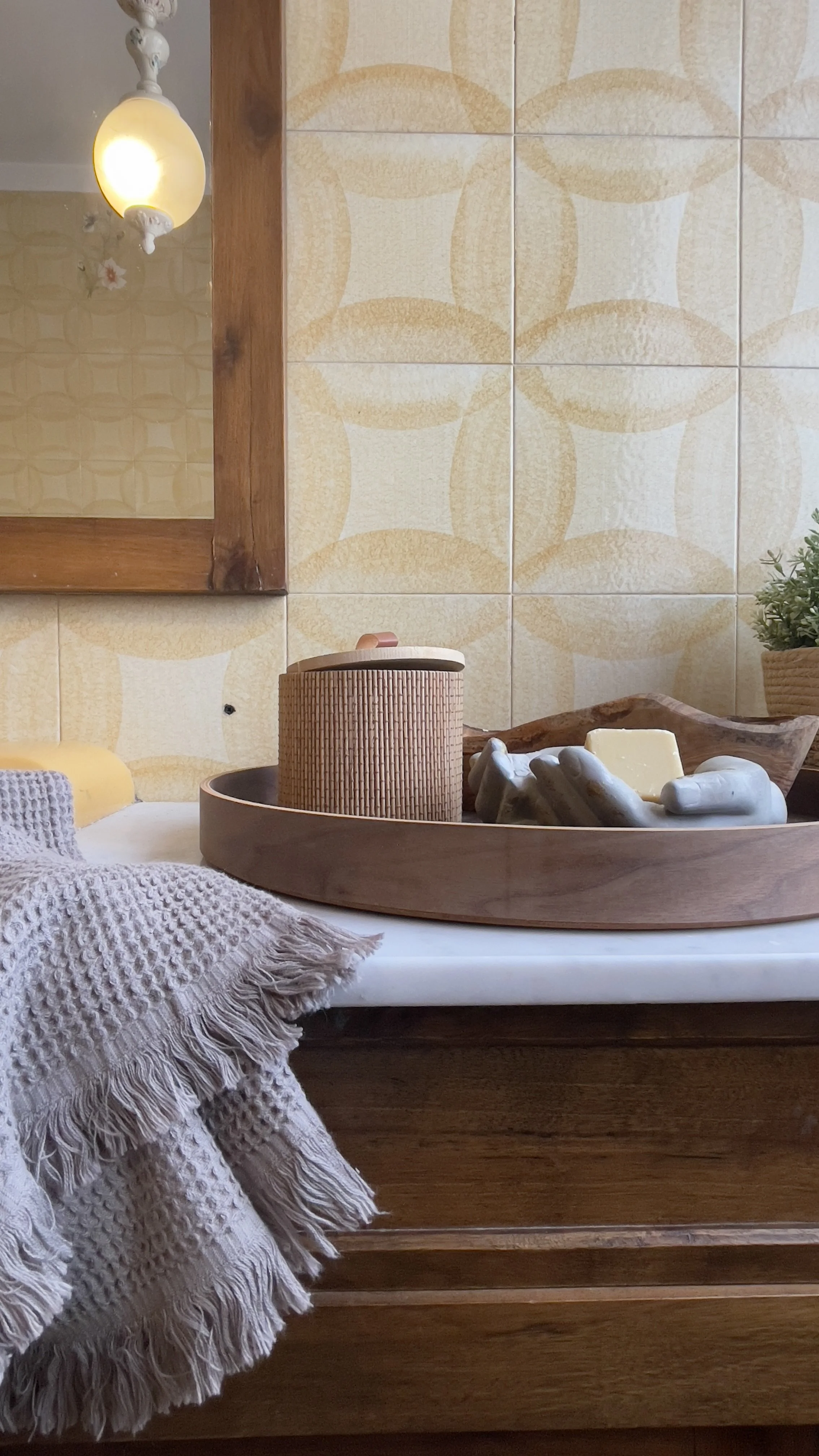 Interno di bagno vintage con piastrelle retrò, lampada sospesa, specchio con cornice in legno, vassoio rotondo con elementi decorativi e asciugamano beige.