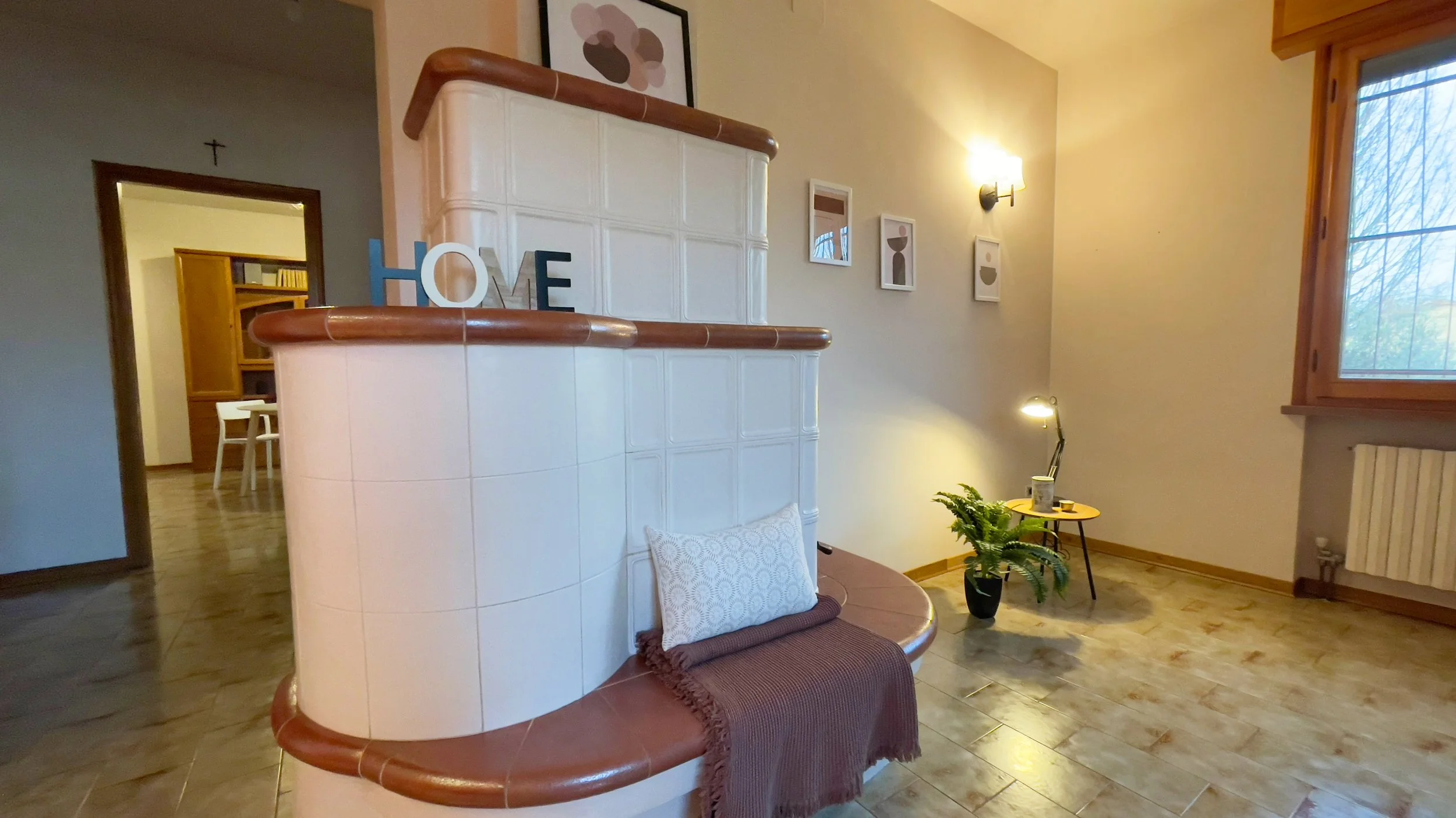 Interno di una casa con una stufa di piastrelle bianche e marroni, decorata con un cuscino e una coperta. Ci sono alcune lettere che formano la parola "HOME" sulla stufa. A destra, un tavolino con una pianta e una lampada. Pareti con quadri e una fin